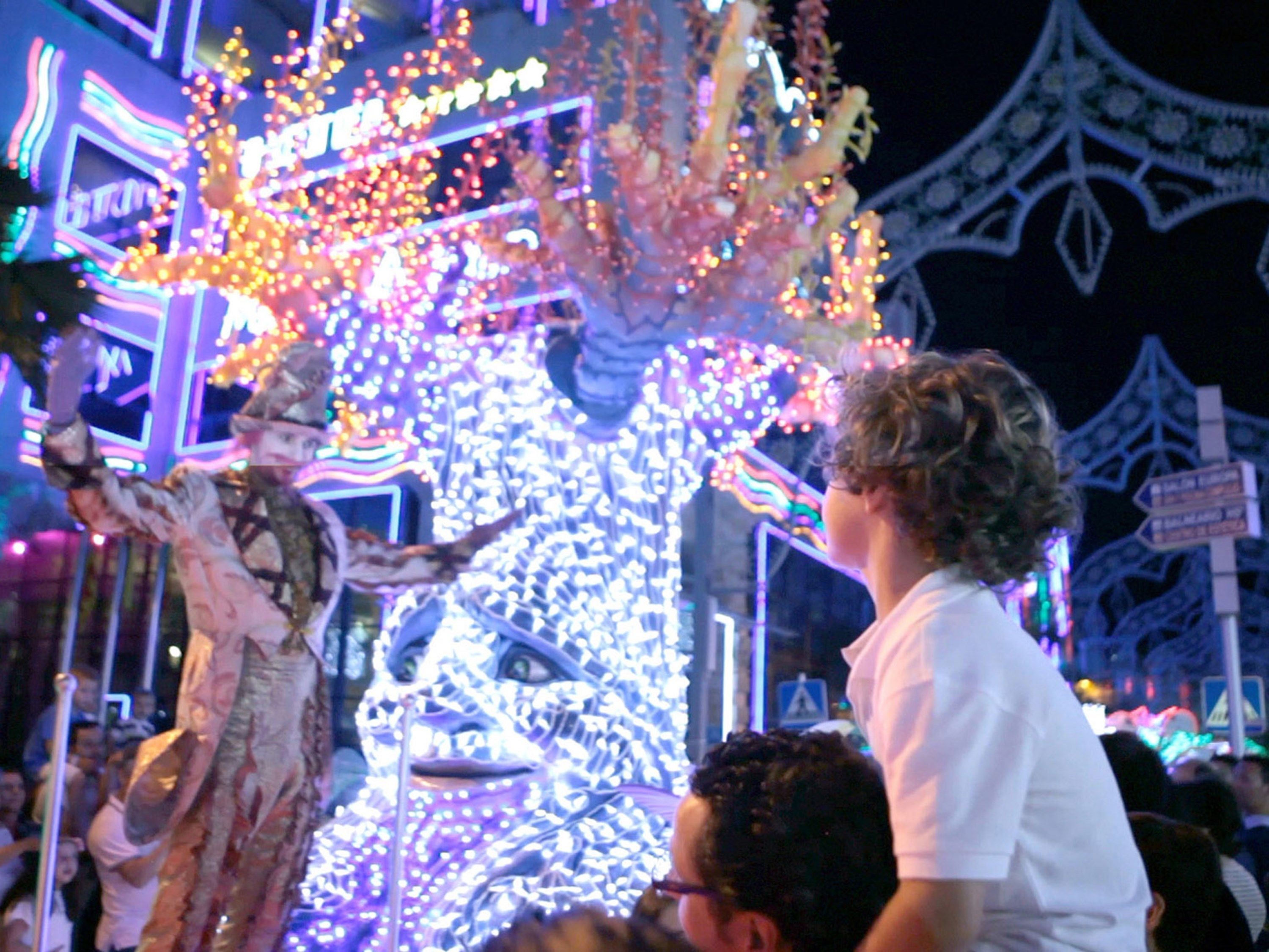 A crowd at a festive event with illuminated structures and a large tree-like decoration.