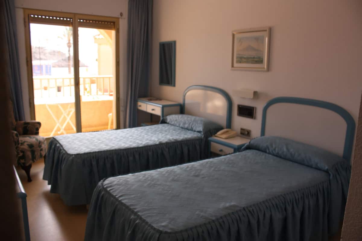A room with two single beds, a desk, a chair, a small table, and a window with curtains.
