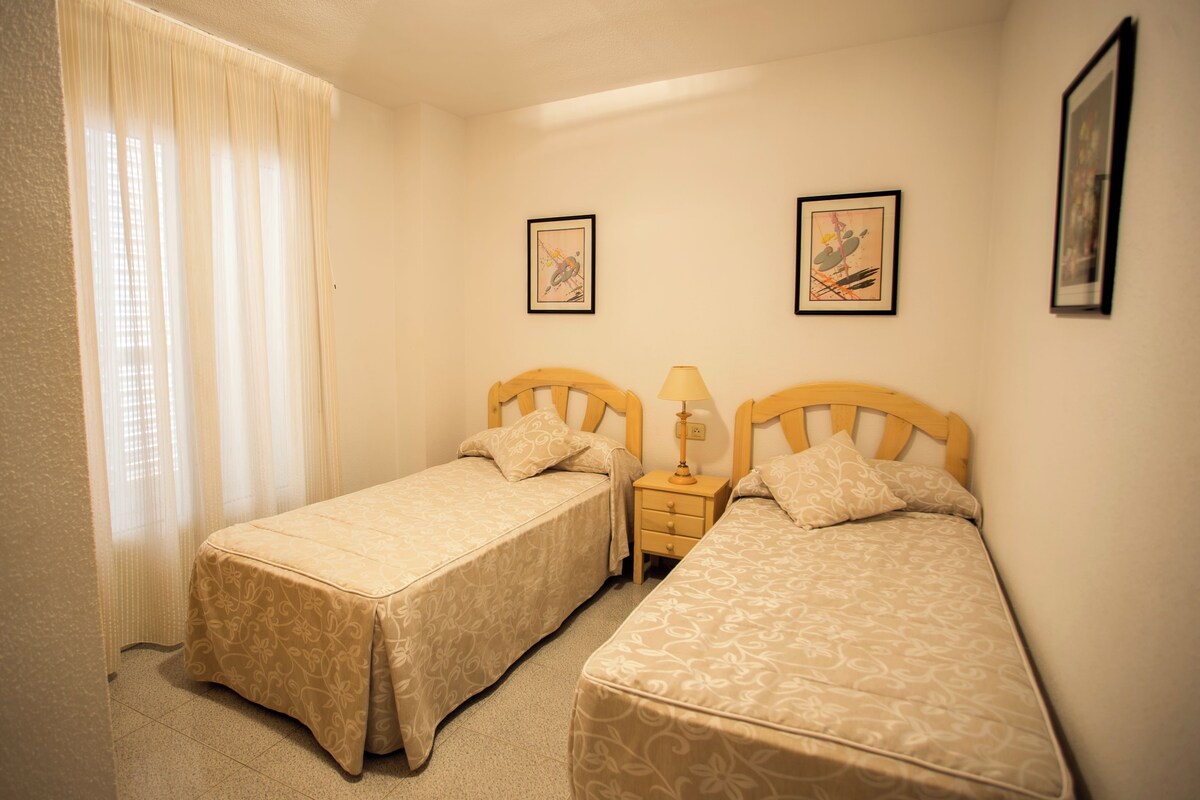 A room with two beds, a nightstand with a lamp, and two framed pictures on the wall.