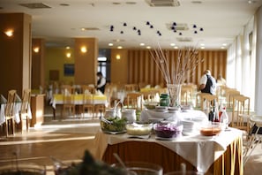 A buffet setup with various food items and serving utensils on a table, surrounded by chairs and tables in a dining area.