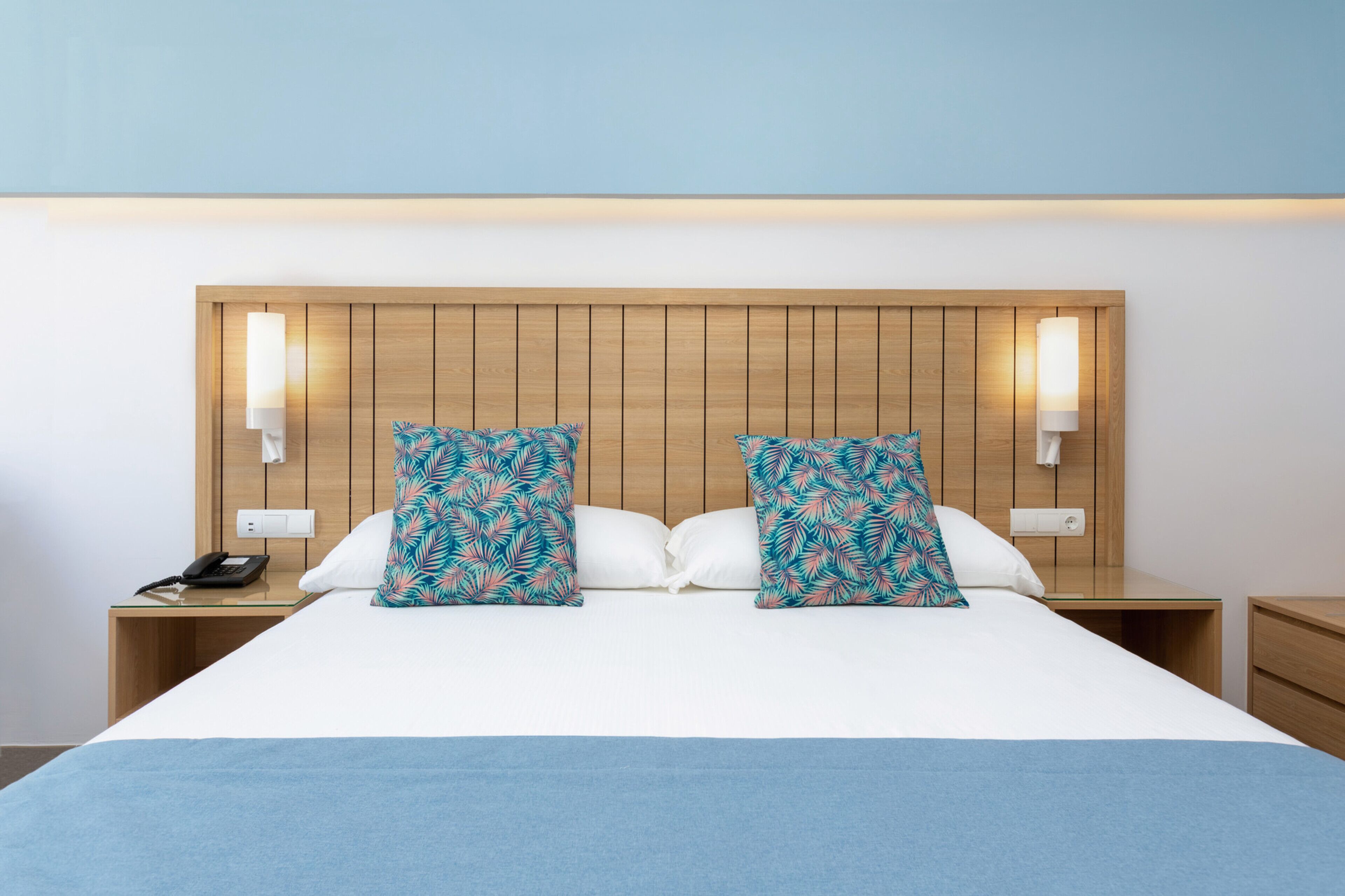 A hotel room with a wooden headboard, two nightstands, a bed with white bedding, and two decorative pillows with a tropical leaf pattern.