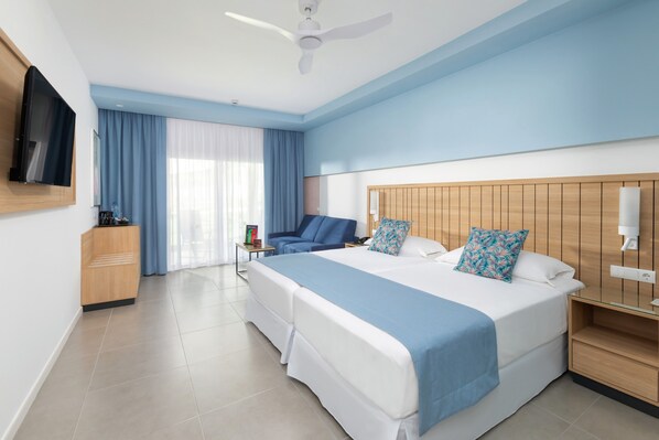 A hotel room with a large bed, a desk, a chair, a TV, and a ceiling fan. - Hotel Riu Palace Palmeras - All Inclusive (San Bartolomé de Tirajana)