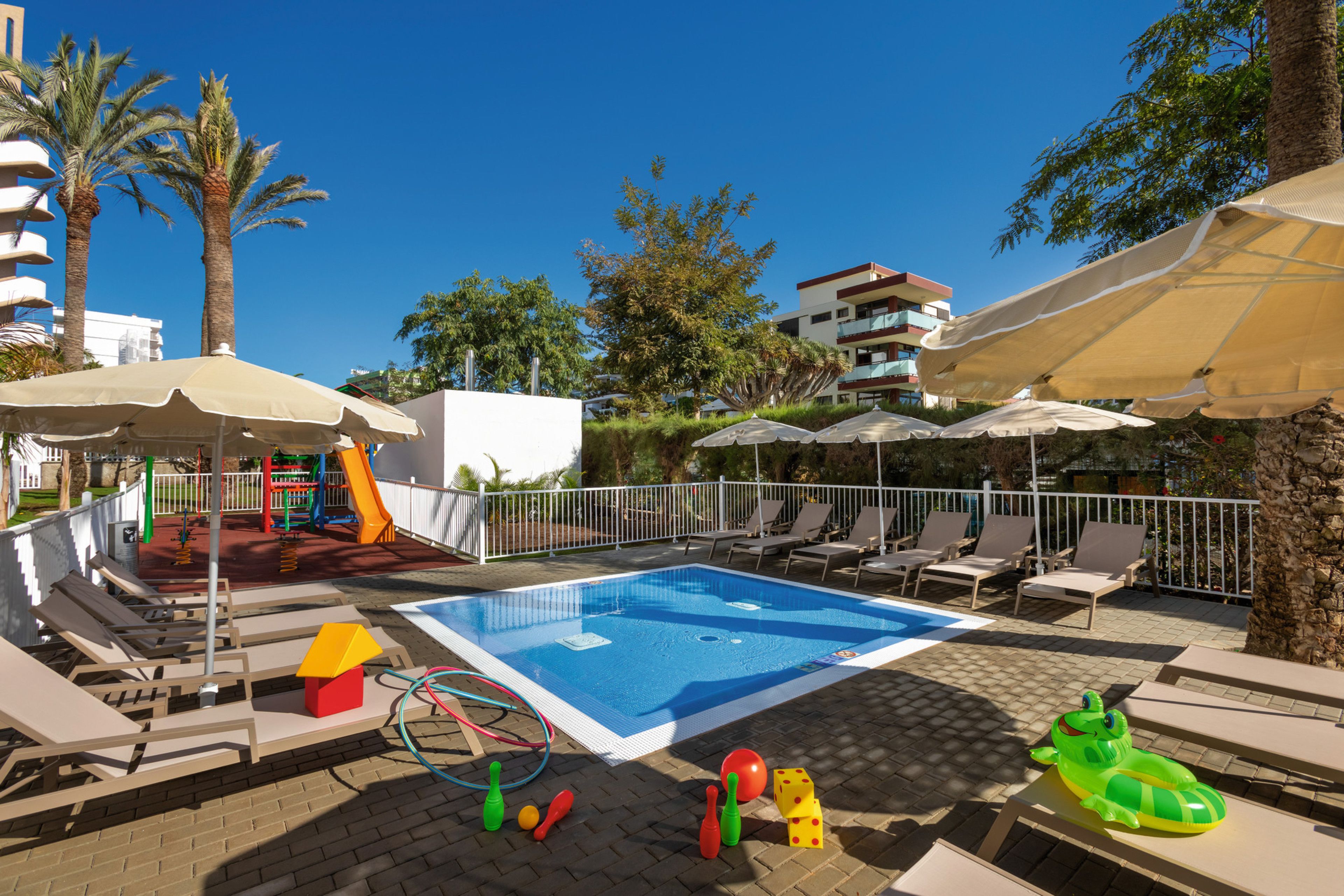 Una piscina con sillas de descanso y sombrillas, rodeada de palmeras y un área de juegos.