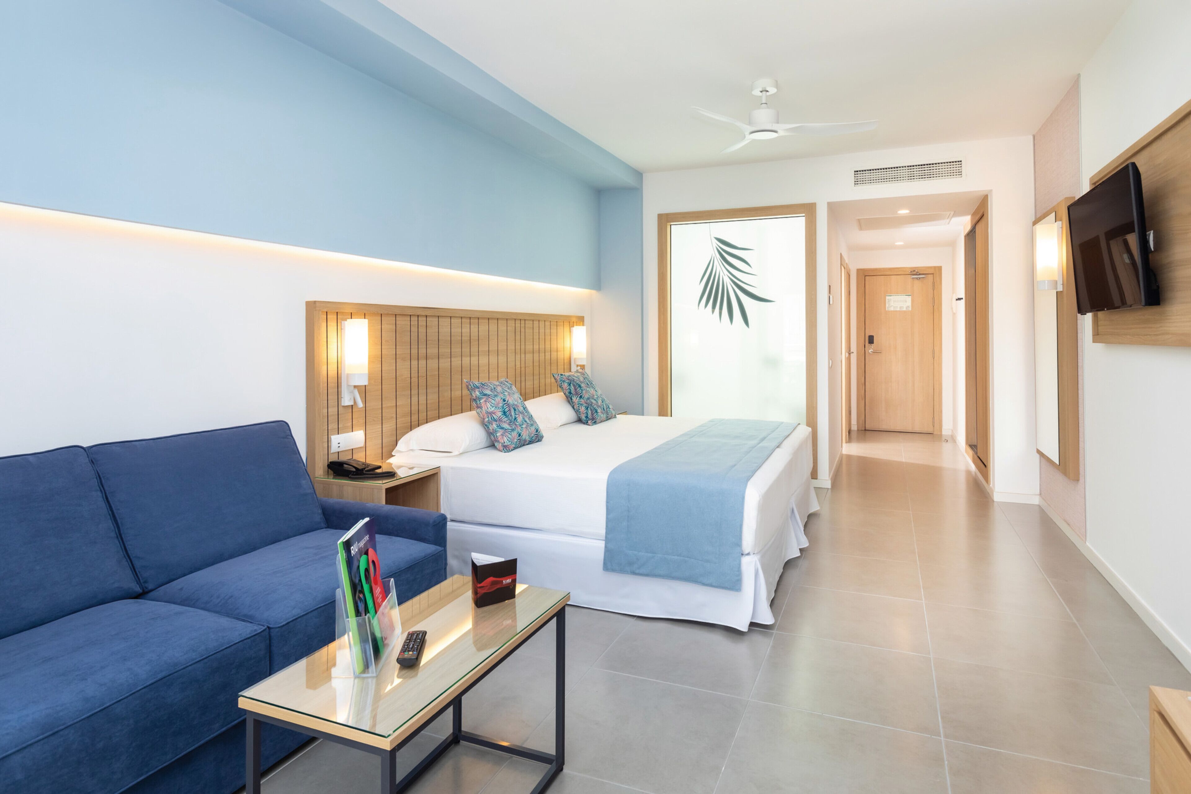Habitación de hotel con una cama, un sofá azul, una mesa de centro de vidrio y un televisor montado en la pared.