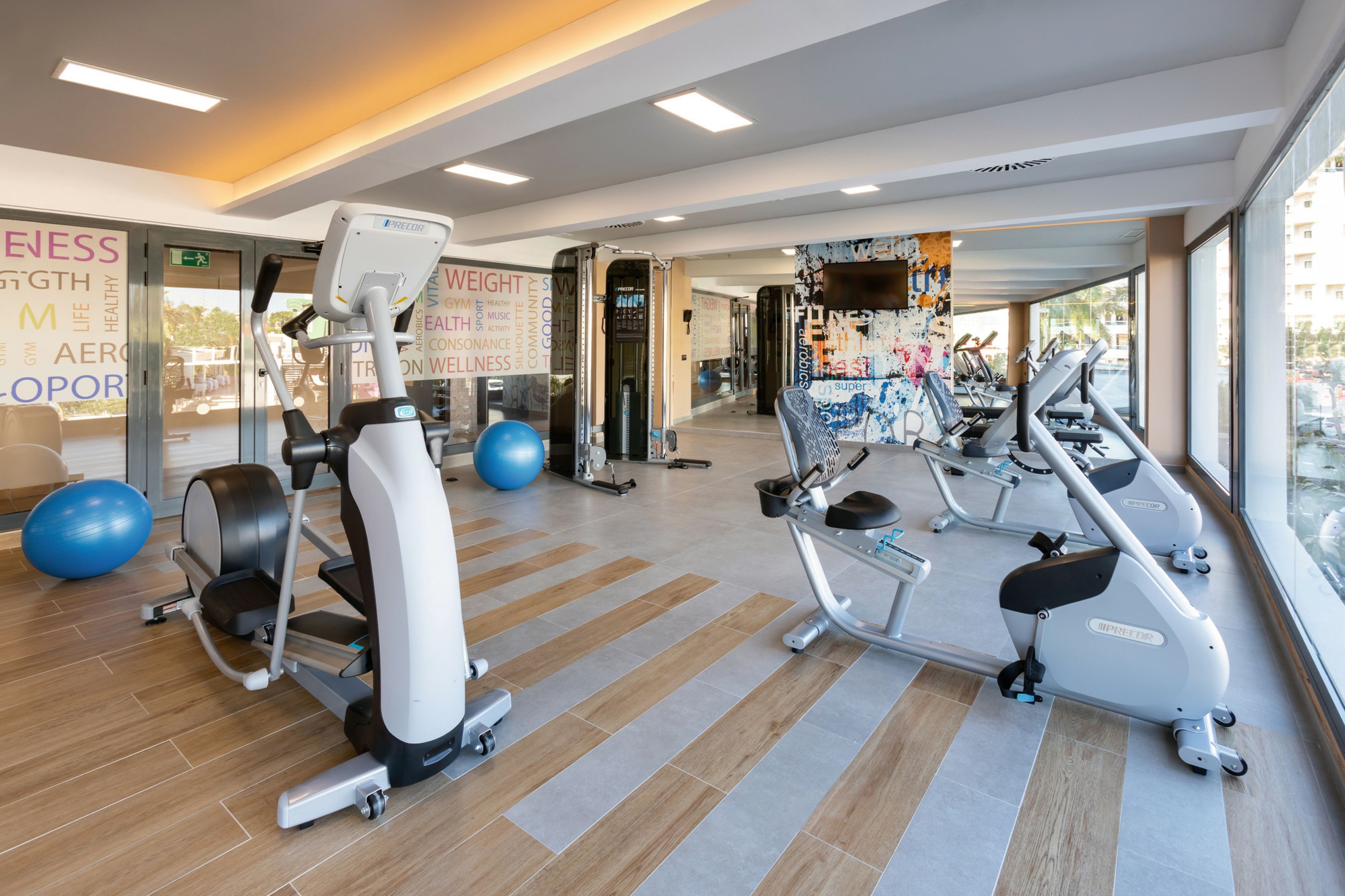 A modern gym with various cardio machines, exercise balls, and a large window offering a view of the outside.