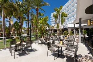 Eine Resort-Terrasse mit Sitzgelegenheiten im Freien, Palmen und einem modernen Gebäude im Hintergrund.