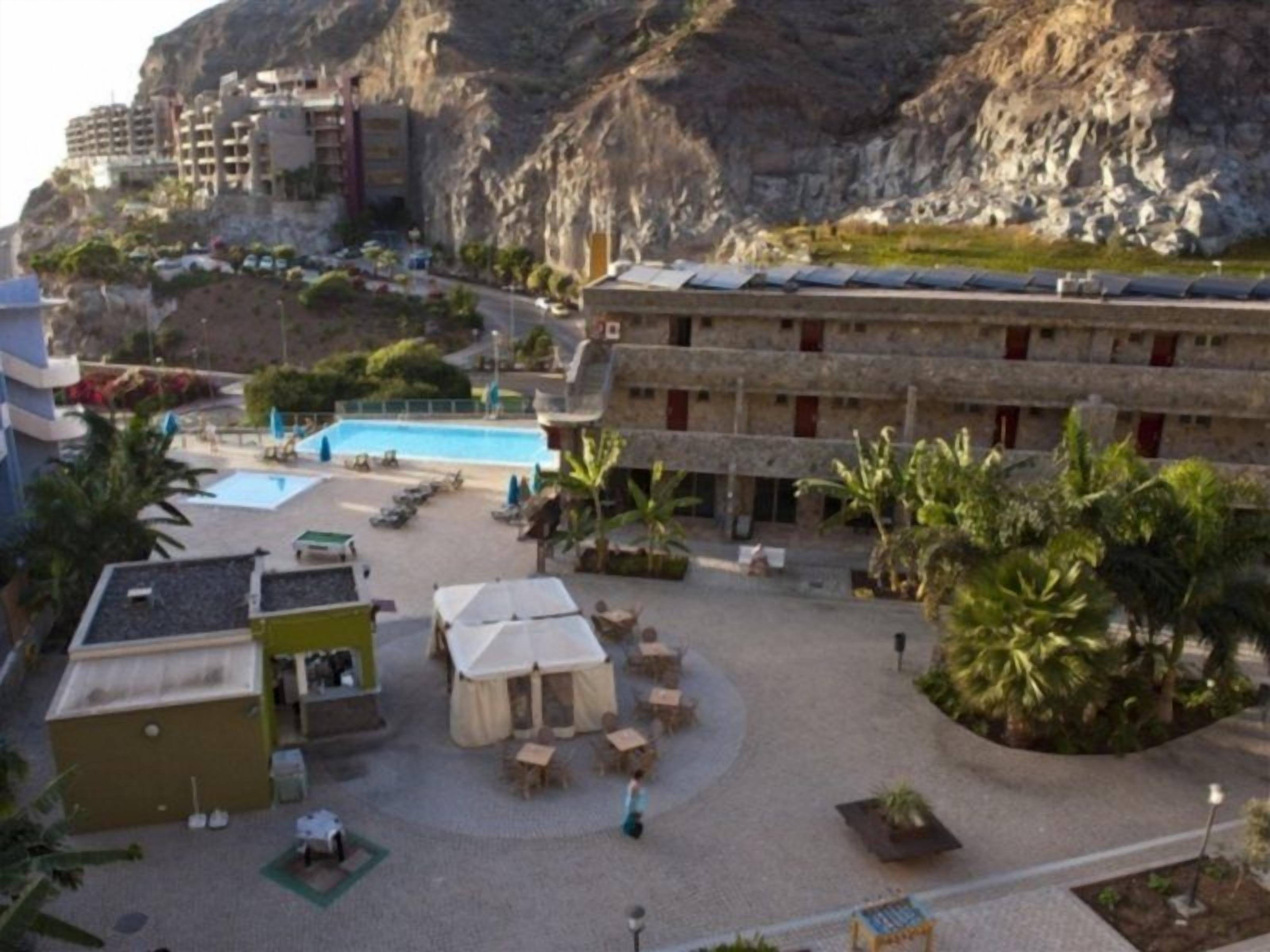 Un patio de hotel con piscina, mobiliario exterior y montañas al fondo.