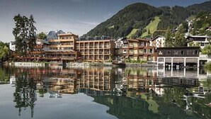 A lakeside hotel with multiple buildings reflecting on the water, surrounded by forested hills. - Seevilla Freiberg (Zell am See)