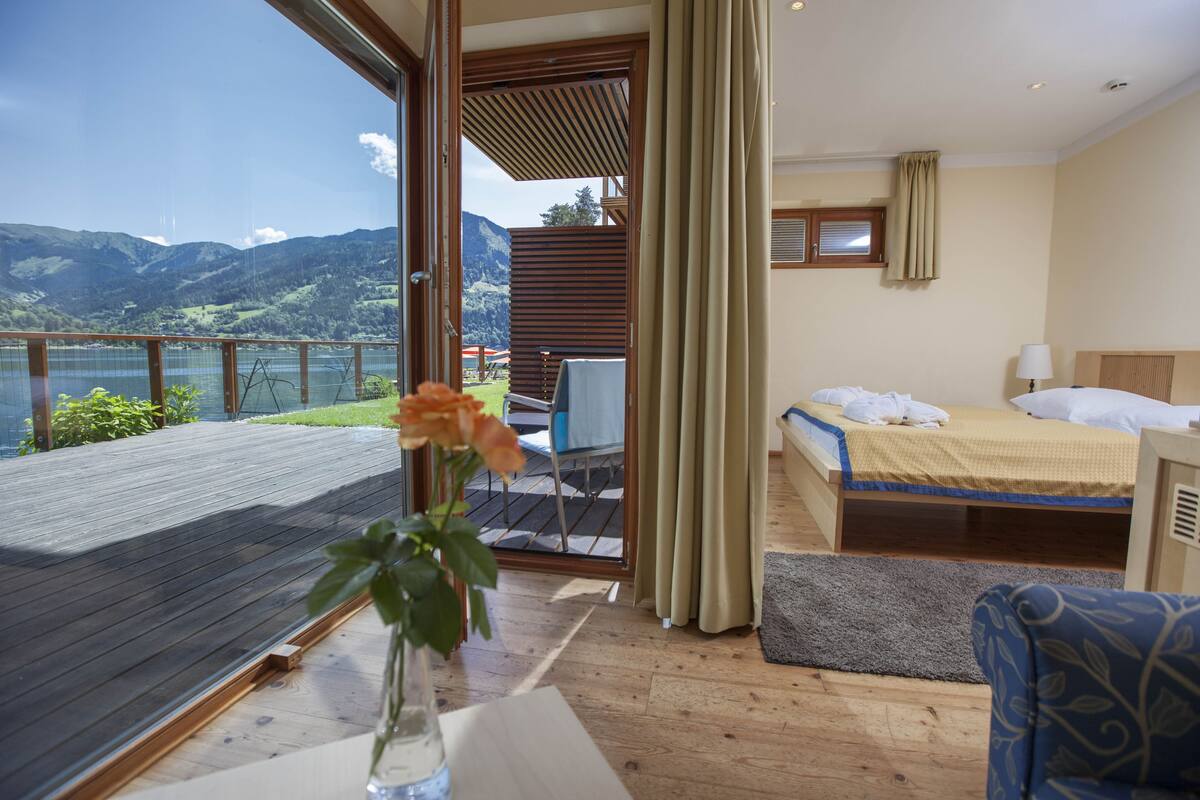A bedroom with a bed, a bedside table, a window with curtains, and a view of mountains and a lake.