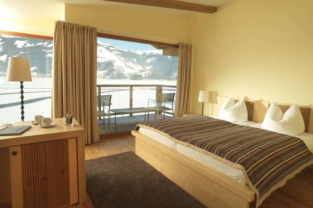 A hotel room with a large bed, a desk with a lamp, and a view of snow-capped mountains through a window.