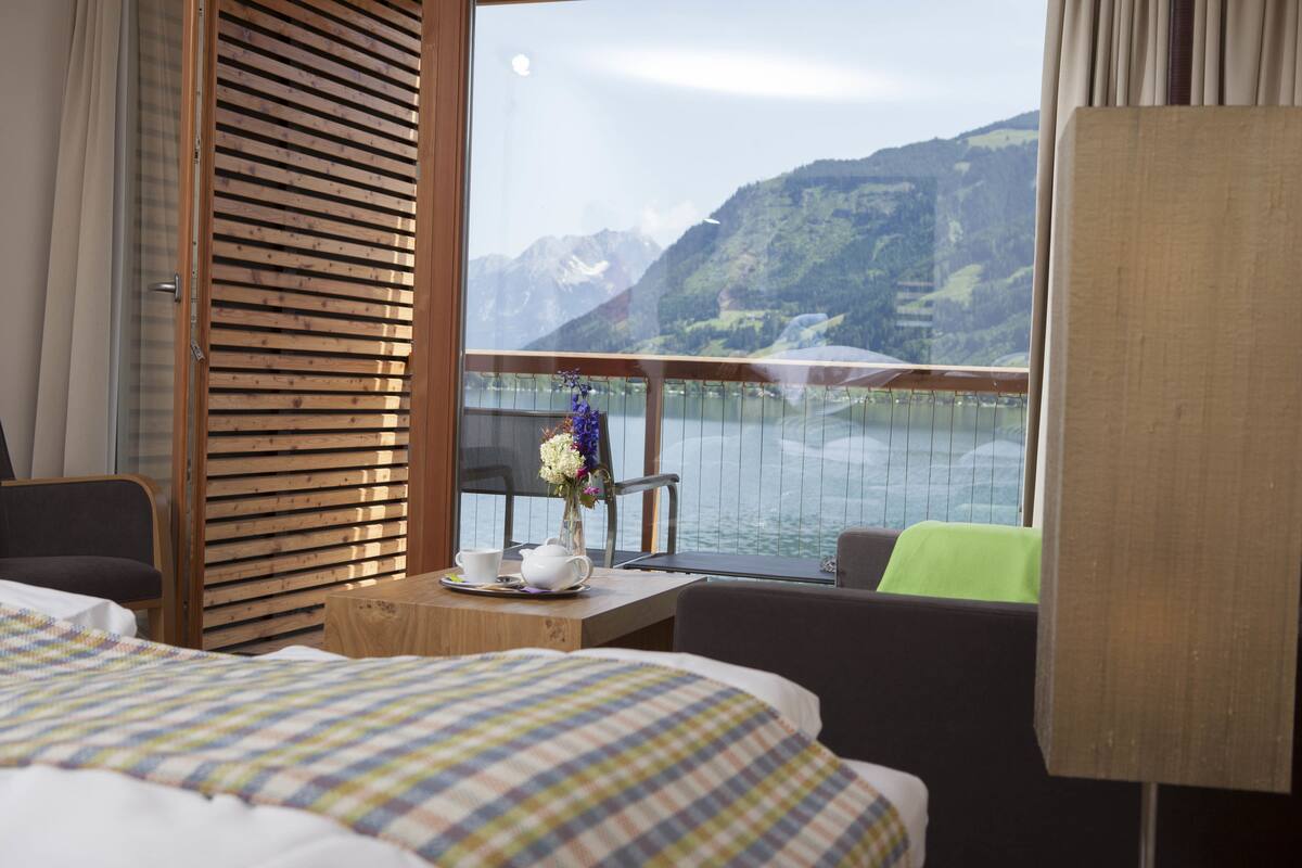 A hotel room with a bed, a small table with a vase of flowers, and a view of a lake and mountains.