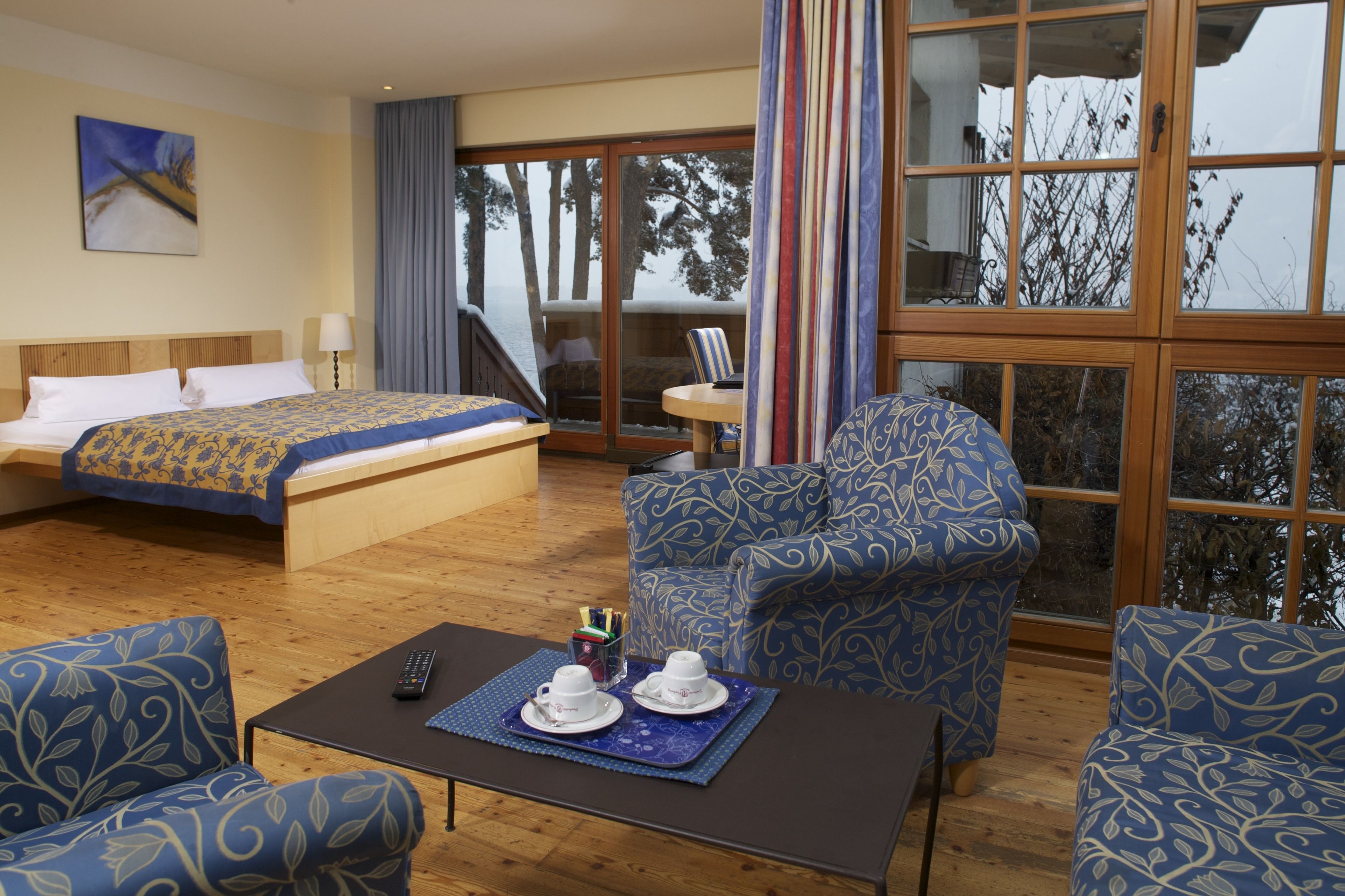 A hotel room with a bed, two blue patterned armchairs, a coffee table with a remote control, and a view of trees through large windows.