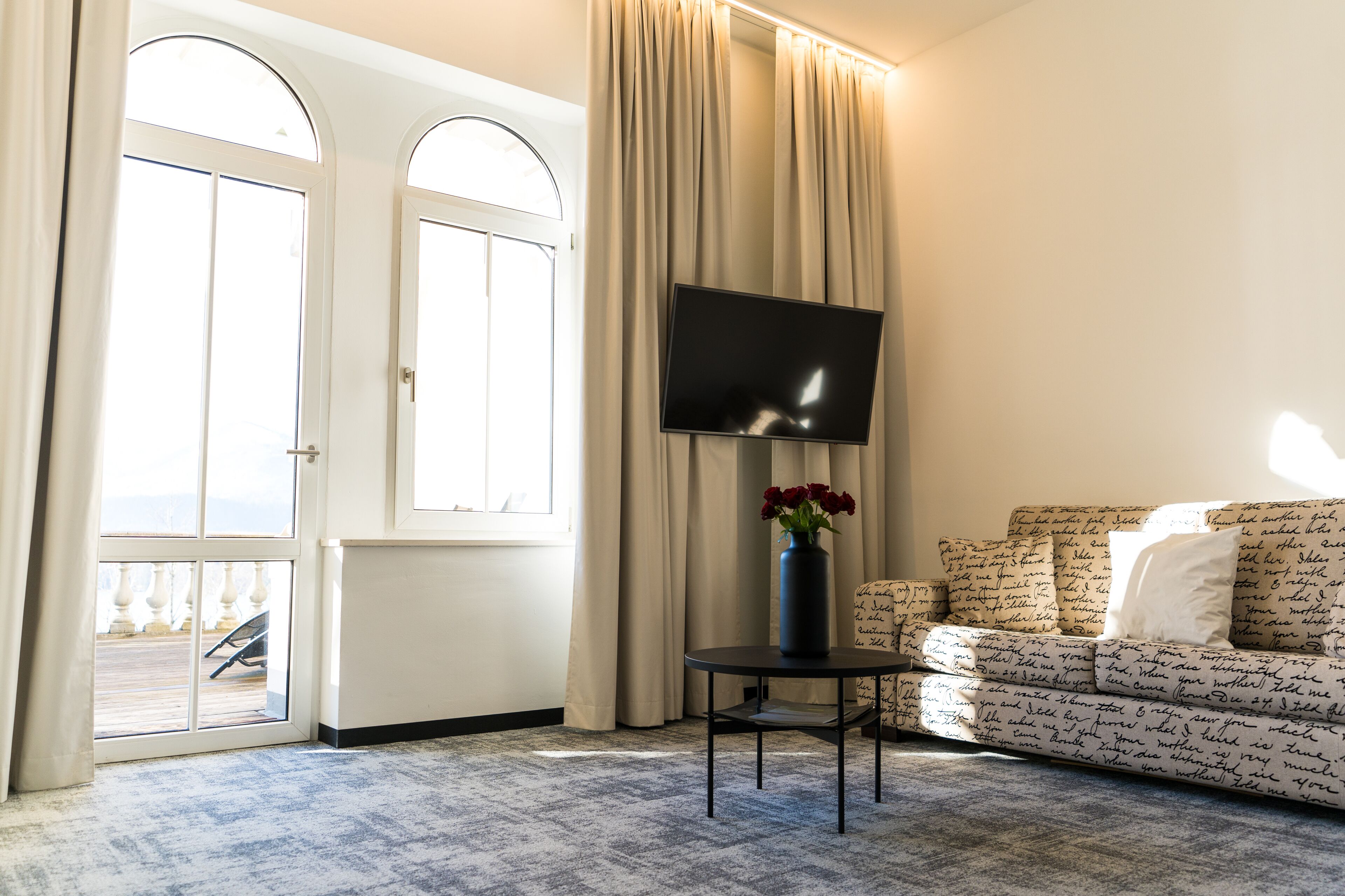 A living room with a large window, a flat-screen TV, a small table with a vase of flowers, and a patterned sofa.