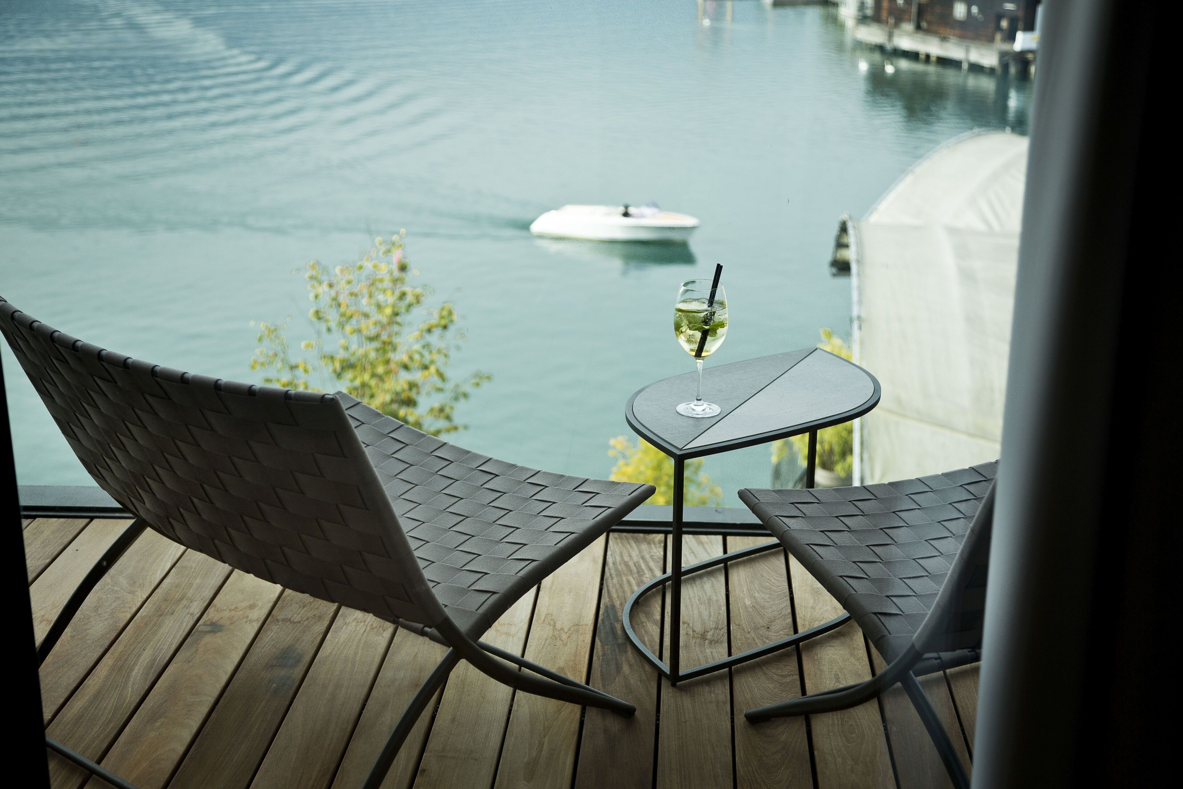 A wooden deck with two metal chairs and a small table, overlooking a calm body of water with a boat and a white tent in the background.