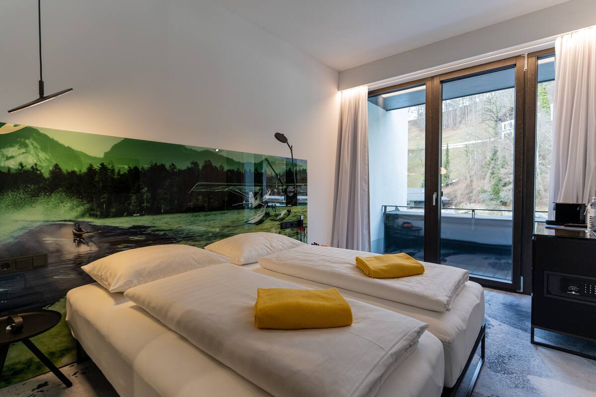 Ein Hotelzimmer mit zwei Betten, einem großen Fenster mit Vorhängen, einem Schreibtisch mit Computer und einem Wandbild einer Landschaft.