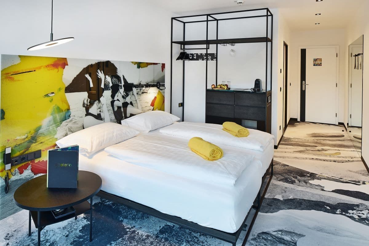 Ein modernes Hotelzimmer mit einem großen Bett, einem Nachttisch, einem schwarzen Schreibtisch, einem Kleiderschrank und einem farbenfrohen abstrakten Gemälde an der Wand.