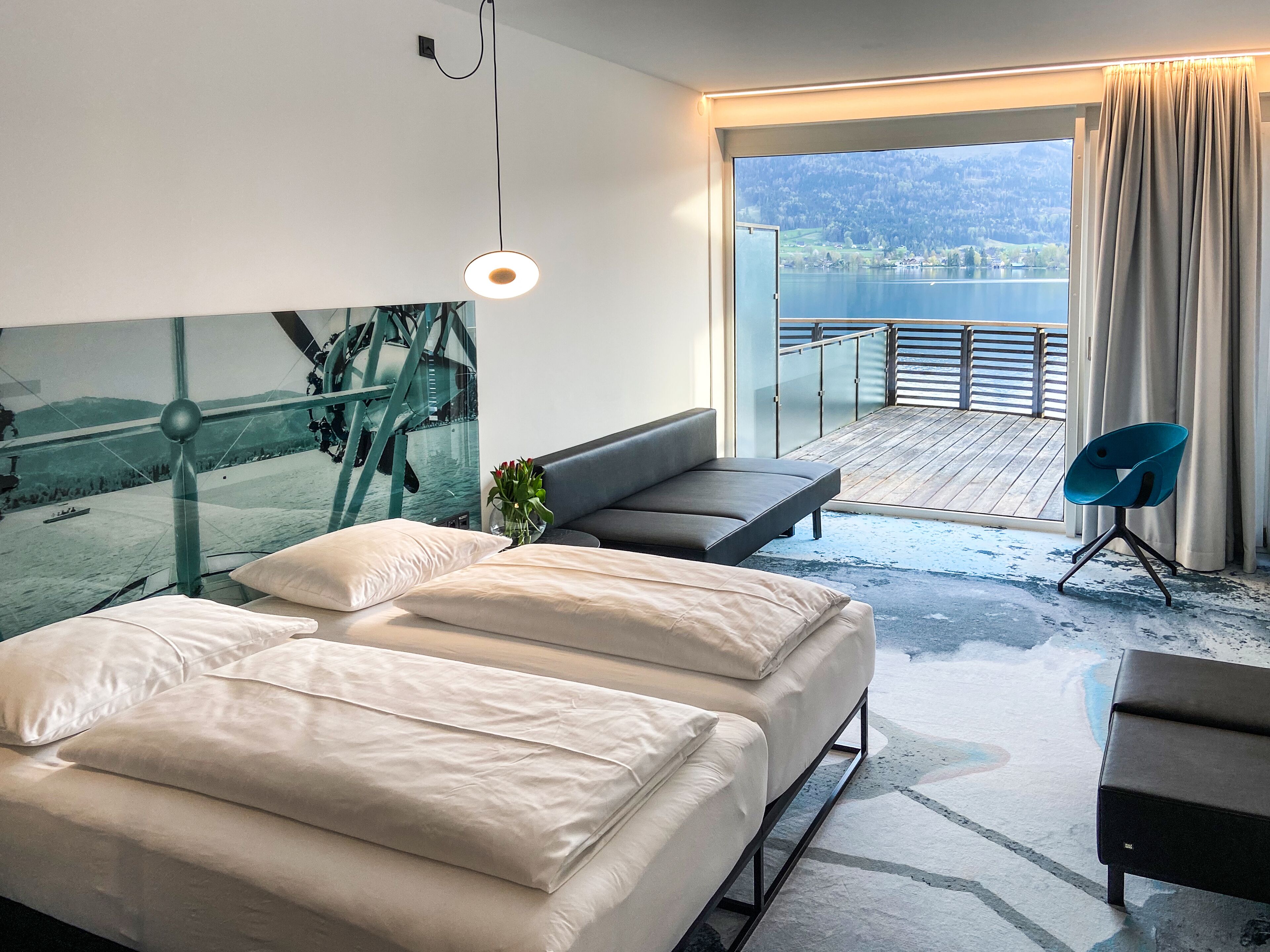 Una habitación de hotel moderna con una cama grande, un sofá, una silla y un balcón con vistas a un lago y montañas.