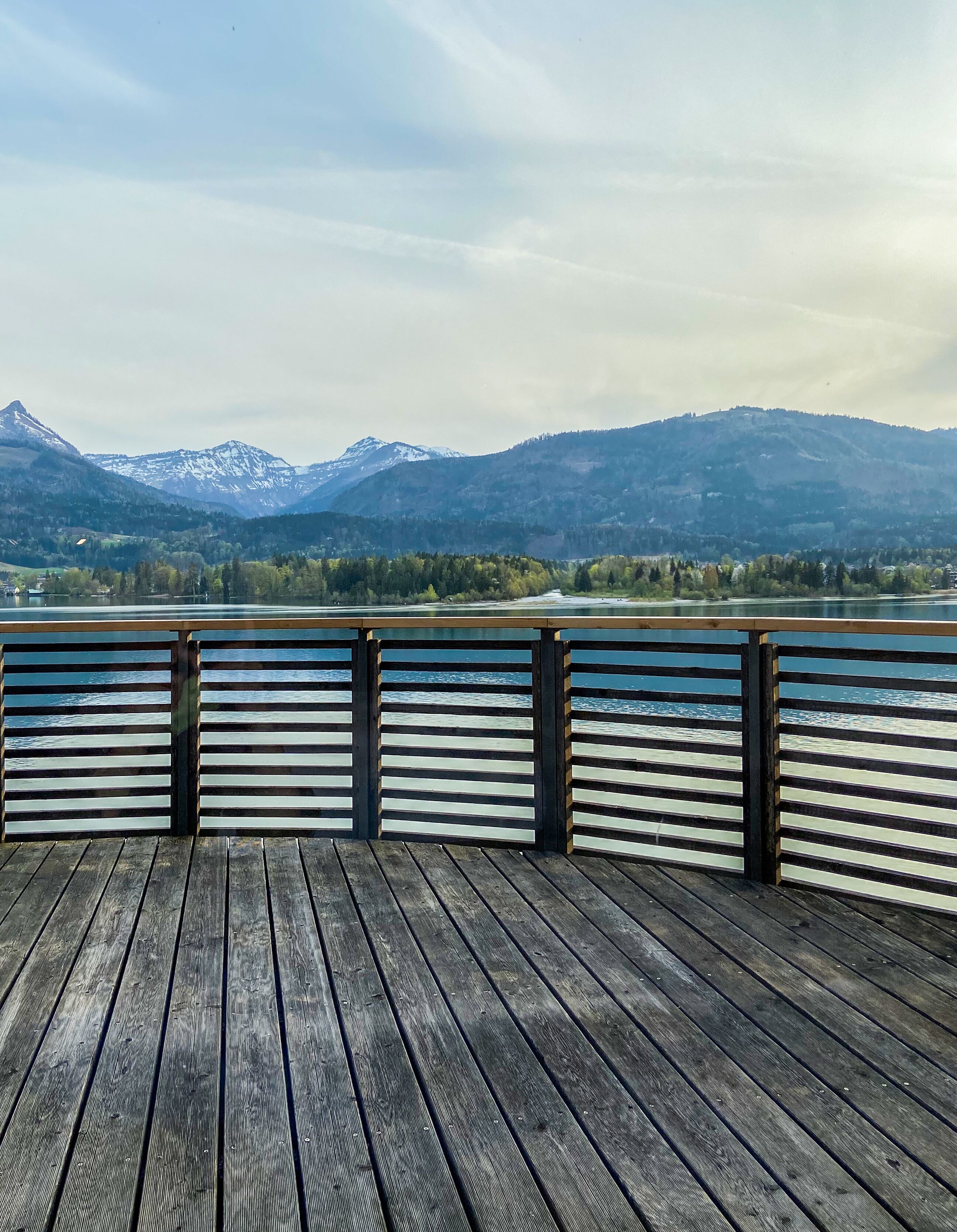 A wooden deck with a railing overlooking a lake and mountains.