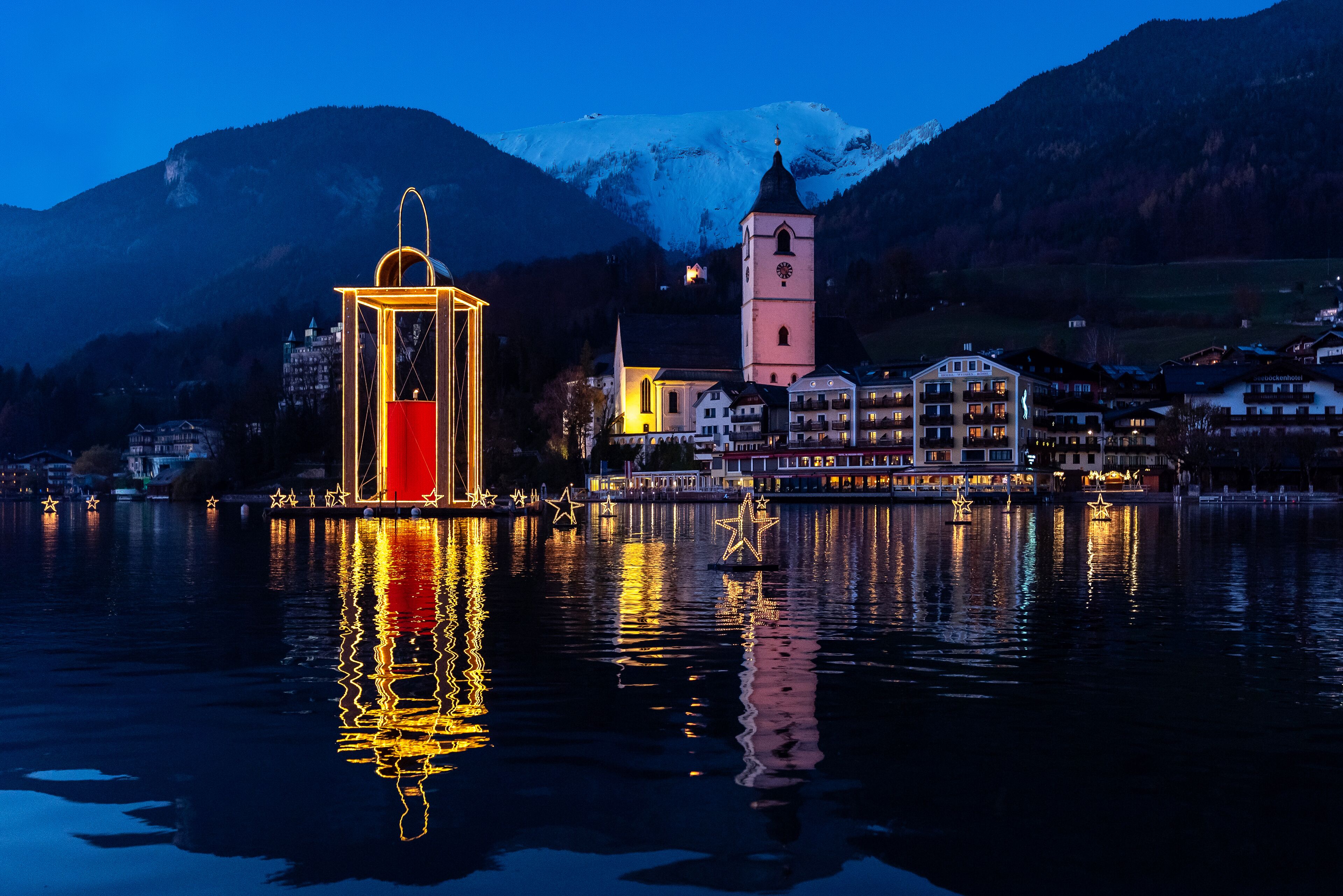 Eine Stadt am Seeufer mit einem markanten Kirchturm, einem beleuchteten Turm und einem Gebäude mit Balkonen mit Blick auf das Wasser.