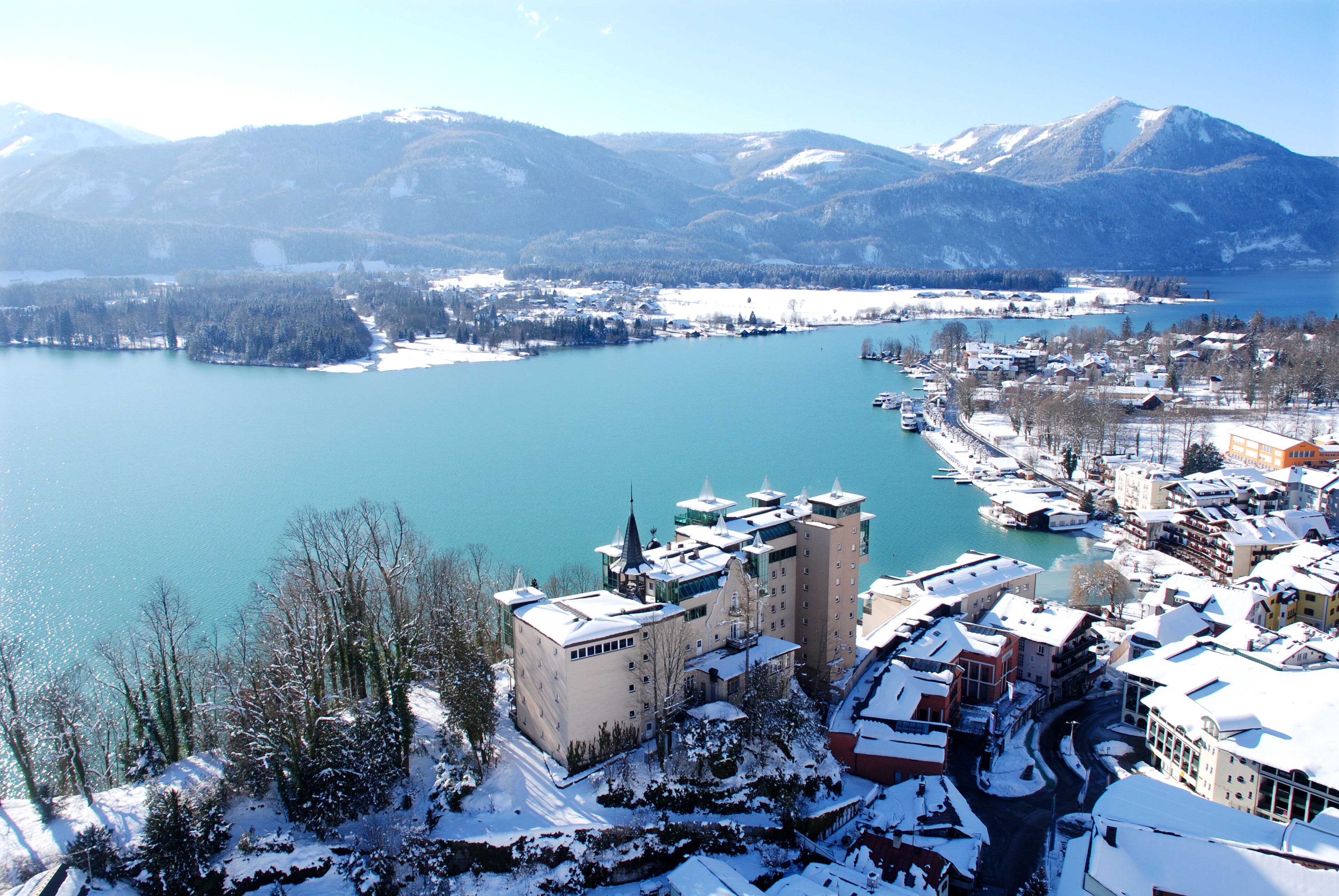 A high-altitude view of a town with a lake, snow-covered buildings, and mountains in the background.