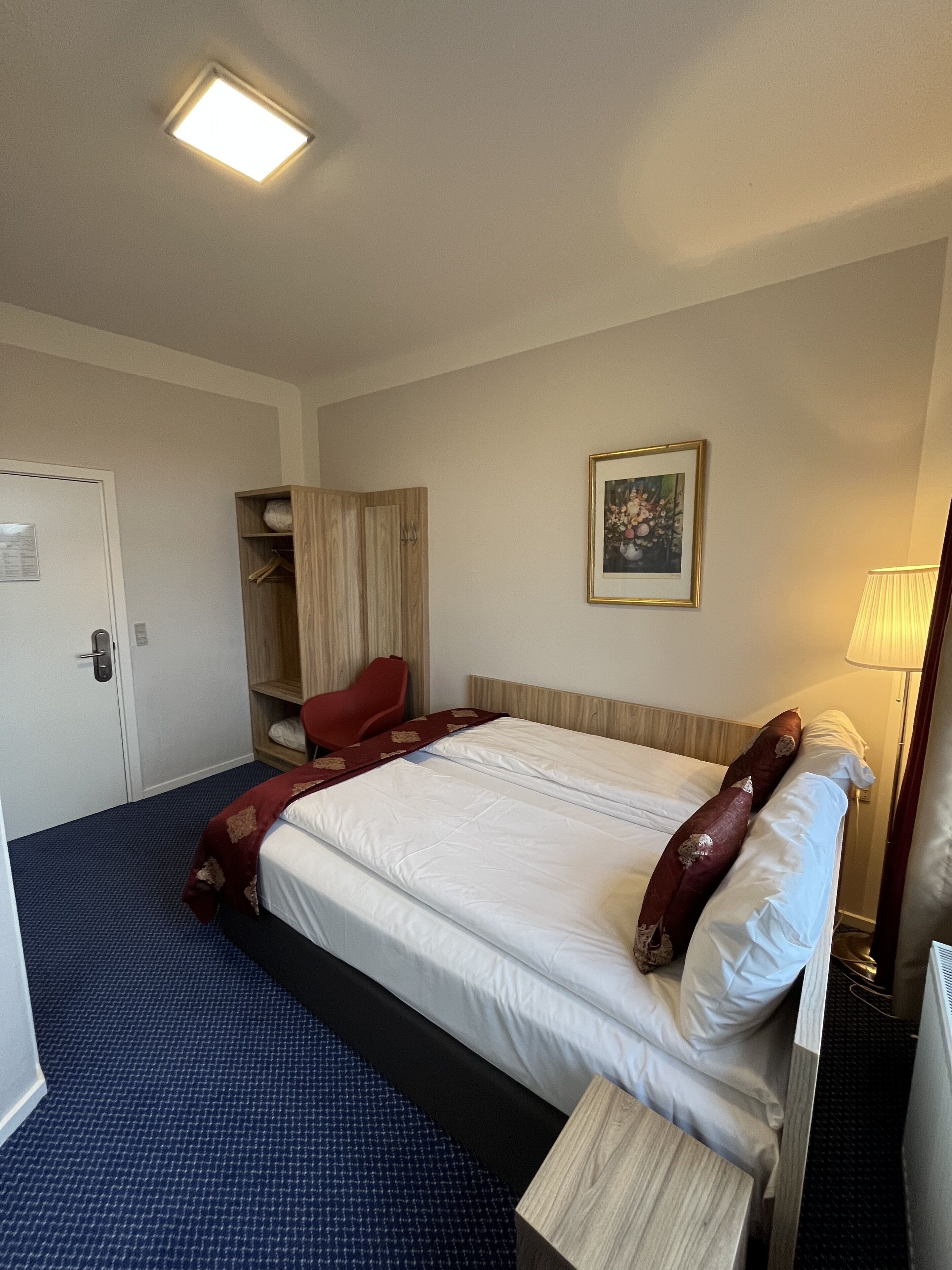 A hotel room with a bed, a red chair, a wardrobe, a framed picture on the wall, and a lamp.