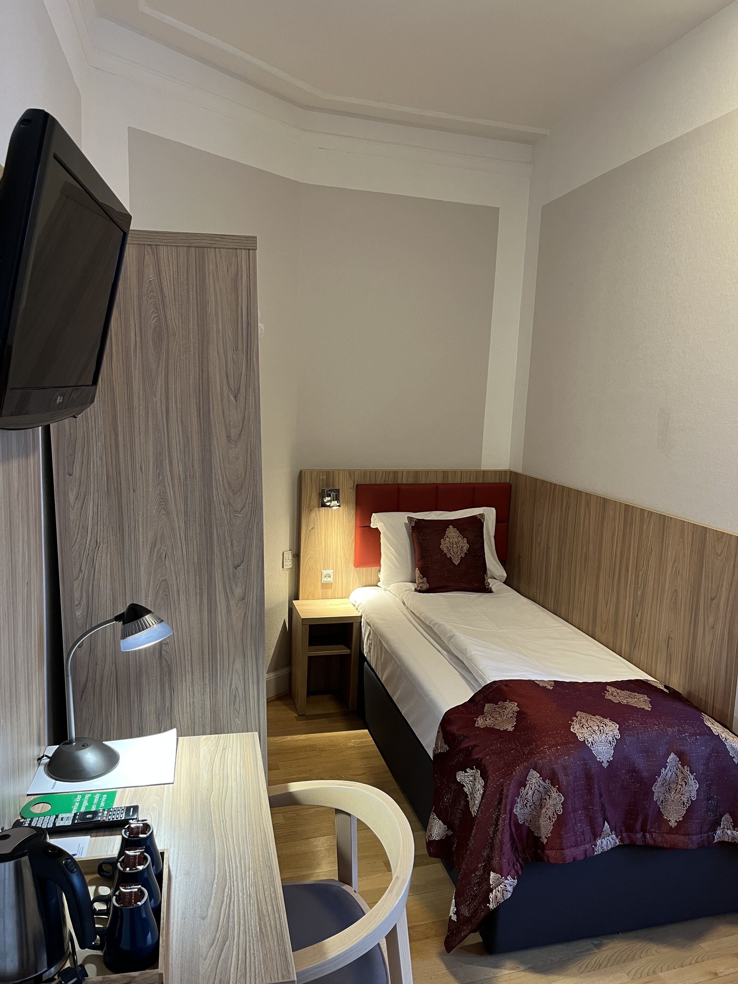 Una camera d'albergo con un letto, una scrivania con lampada, una sedia e un televisore fissato alla parete.