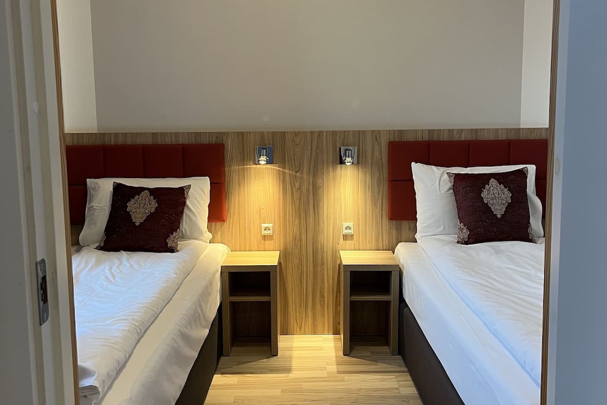 Hotellihuone, jossa on kaksi sänkyä, puiset päätylevyt, yöpöydät ja seinään kiinnitetyt valaisimet.