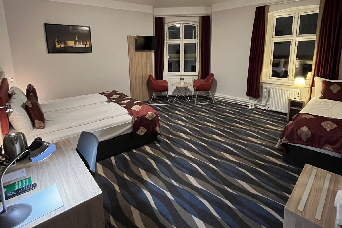Hotellihuone, jossa on sänky, työpöytä tietokoneella, tuoli, pieni pöytä ja ikkuna, jossa on verhot.