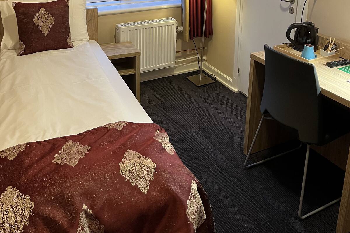 Hotellihuone, jossa on sänky, työpöytä, patteri, ikkuna verhoilla ja lamppu.
