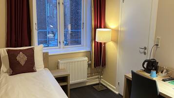 A hotel room with a bed, a desk, a radiator, a window with curtains, and a lamp.