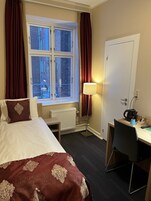 Una habitación de hotel con cama, escritorio, radiador, ventana con cortinas y lámpara.