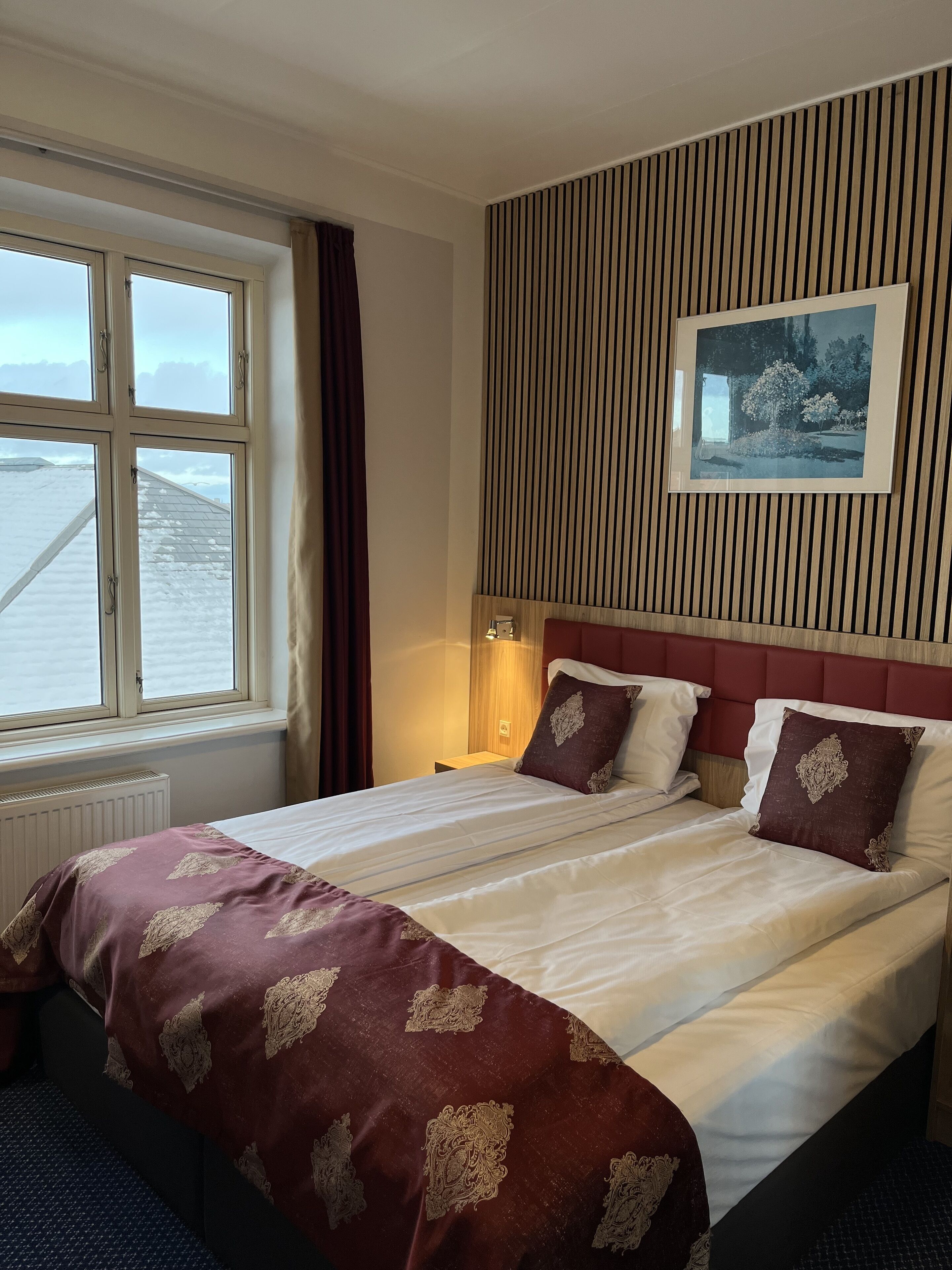 Ein Hotelzimmer mit einem großen Bett, einem Fenster mit Blick auf verschneite Dächer, einer Holzwand und einem gerahmten Bild.