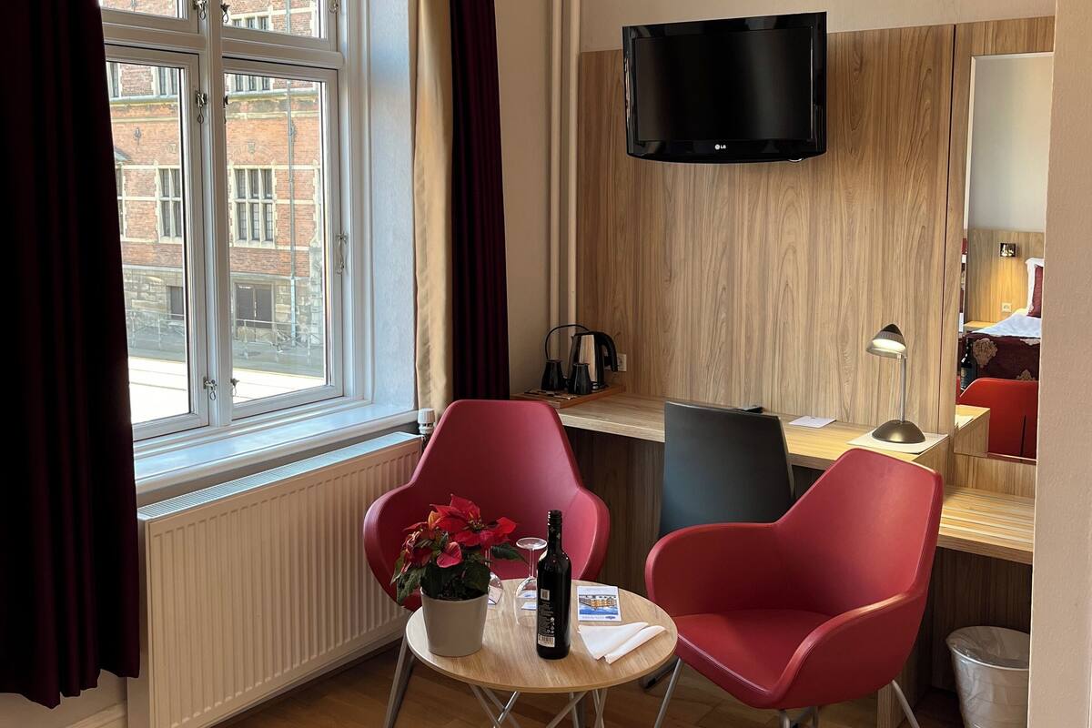 Moderni hotellihuone, jossa on suuri ikkuna, punaiset tuolit, pieni pöytä, jonka päällä on pullo ja lasi, seinälle asennettu televisio ja puulattia.