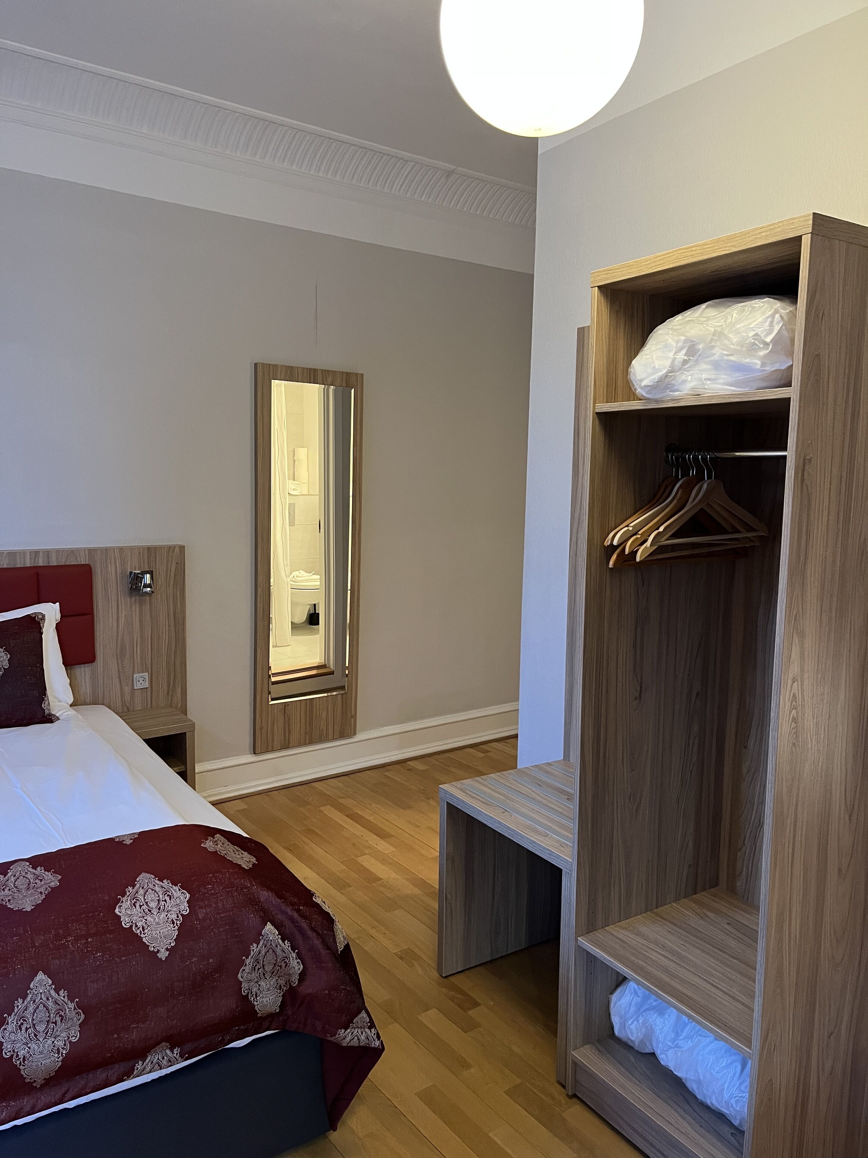 A hotel room with a bed, a wooden wardrobe, a bench, and a mirror.
