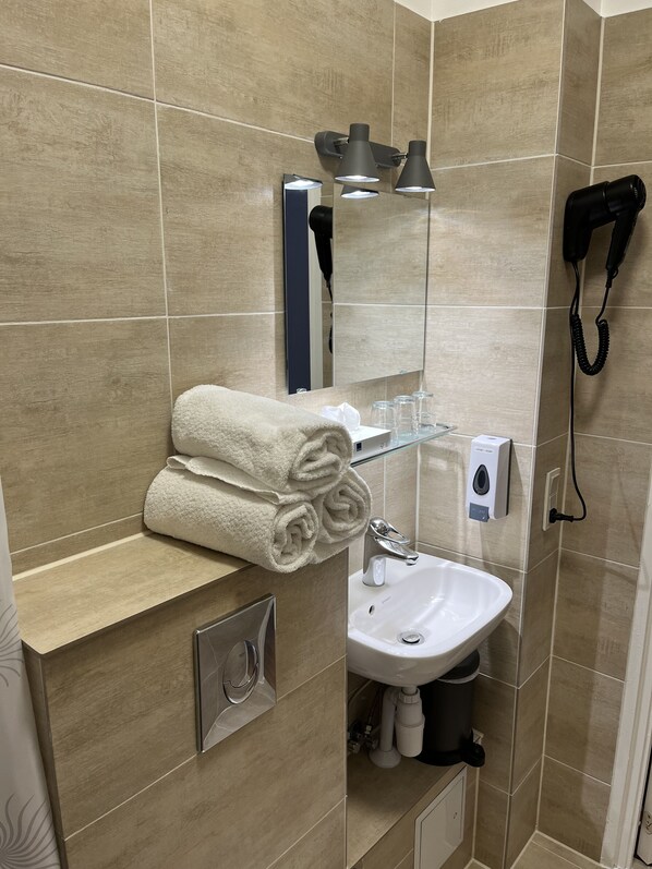 Un baño con azulejos color beige, un lavabo, un espejo, un perchero para toallas, un secador de pelo y un dispensador de jabón.