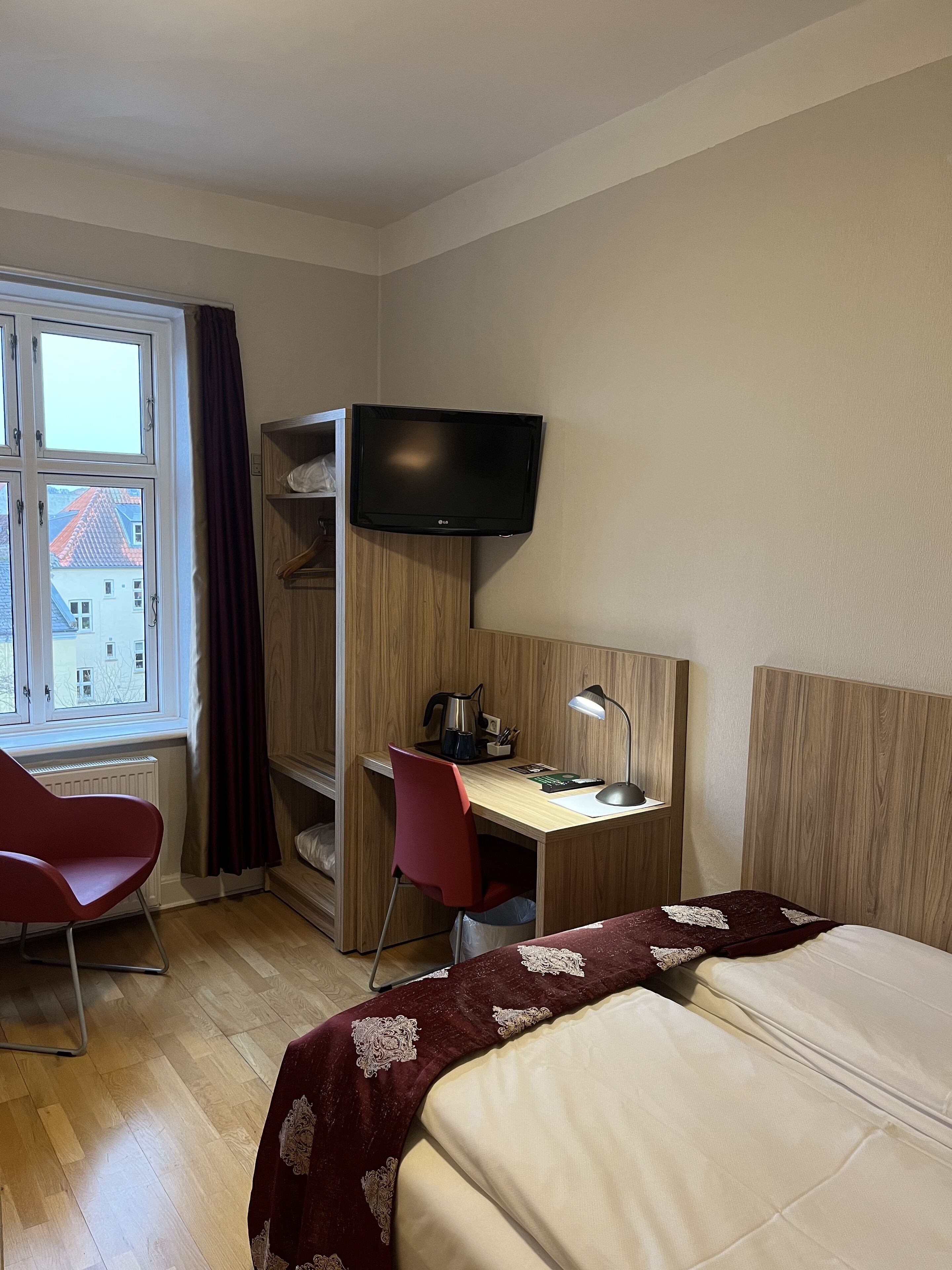 A hotel room with a bed, a desk with a computer, a TV mounted on the wall, a window with curtains, and a red chair.