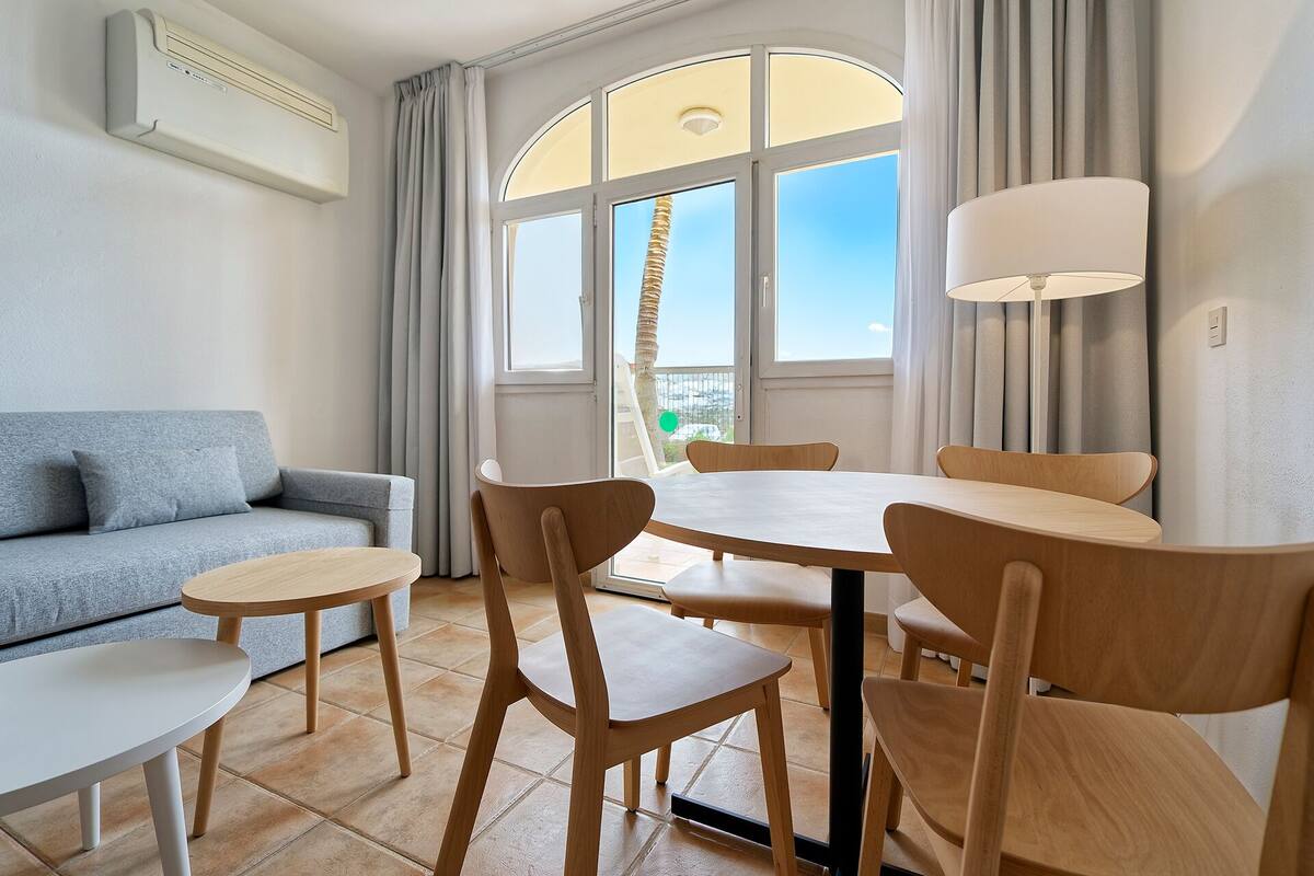 A modern living room with a grey sofa, a round dining table with wooden chairs, and a large window with curtains.
