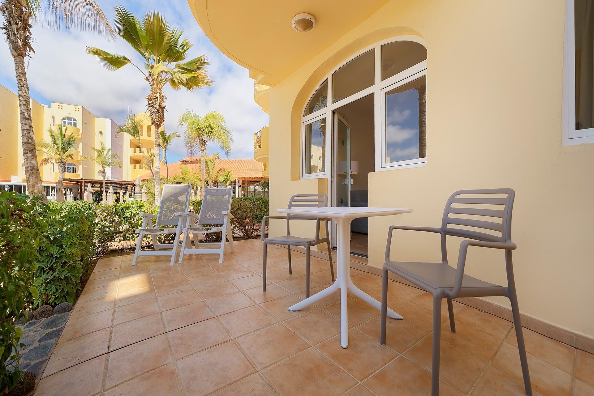 An outdoor patio with a table and chairs, palm trees, and a building with arched windows.