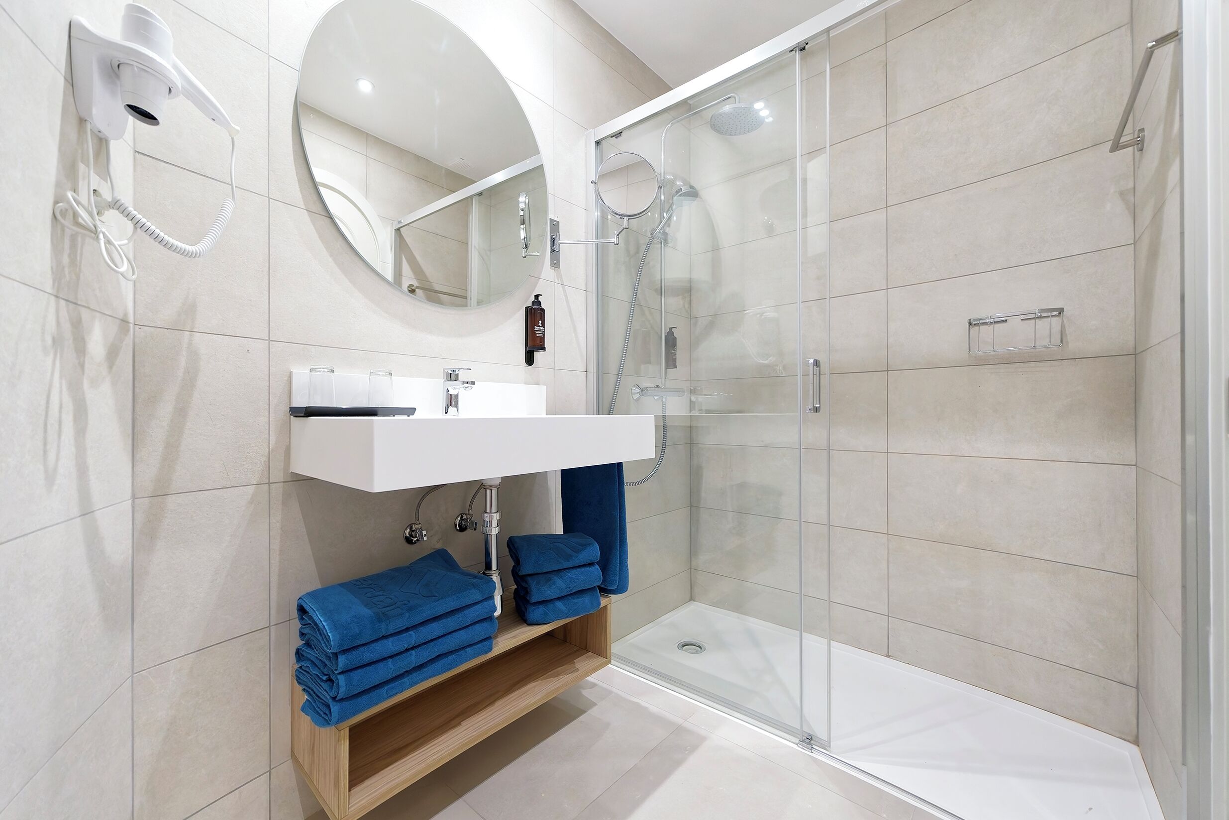 Un baño moderno con cabina de ducha de vidrio, un lavamanos blanco con espejo y toallas azules sobre una estantería de madera.