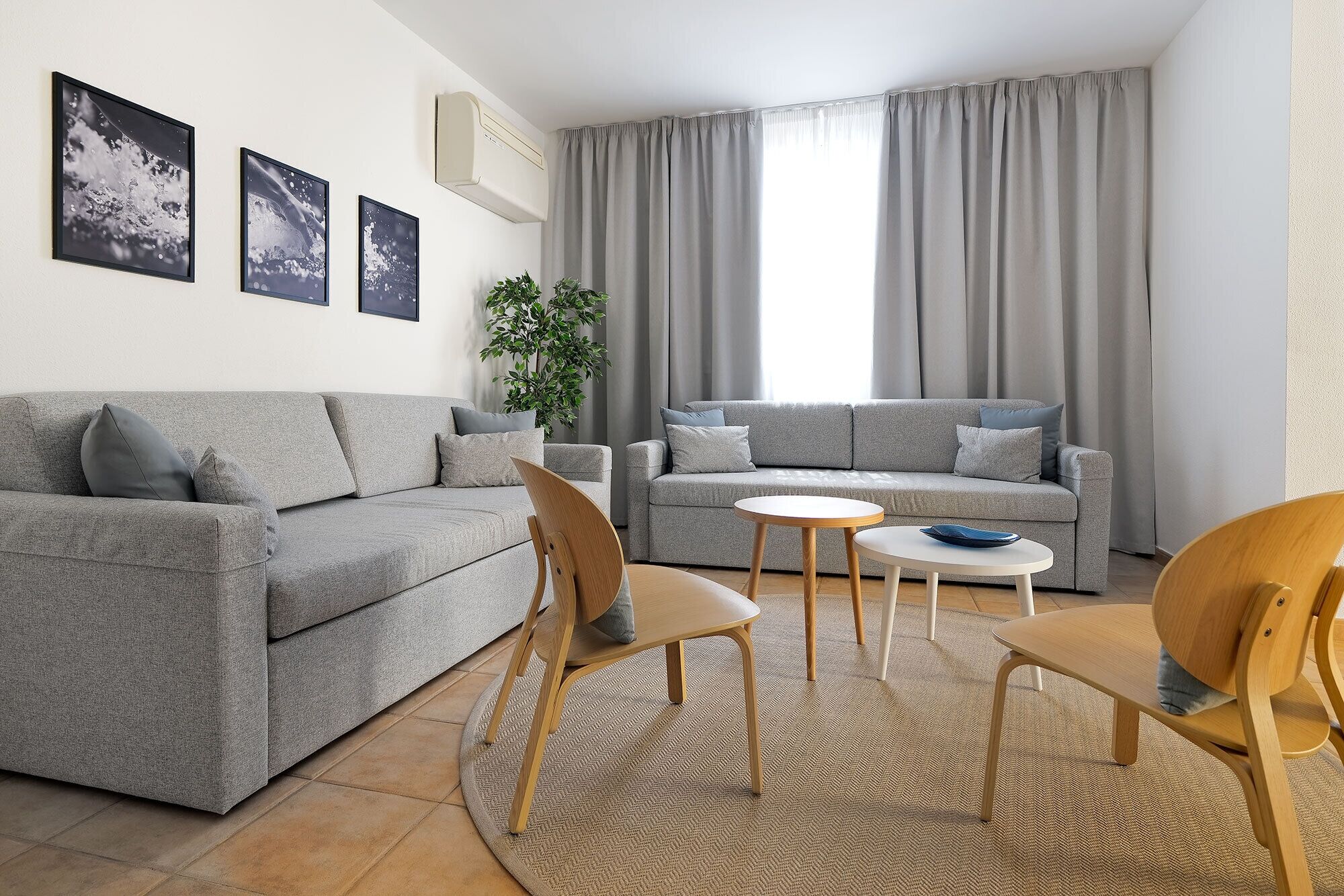 Un salon moderne avec un canapé gris, deux fauteuils, une petite table ronde et une grande fenêtre agrémentée de rideaux gris.