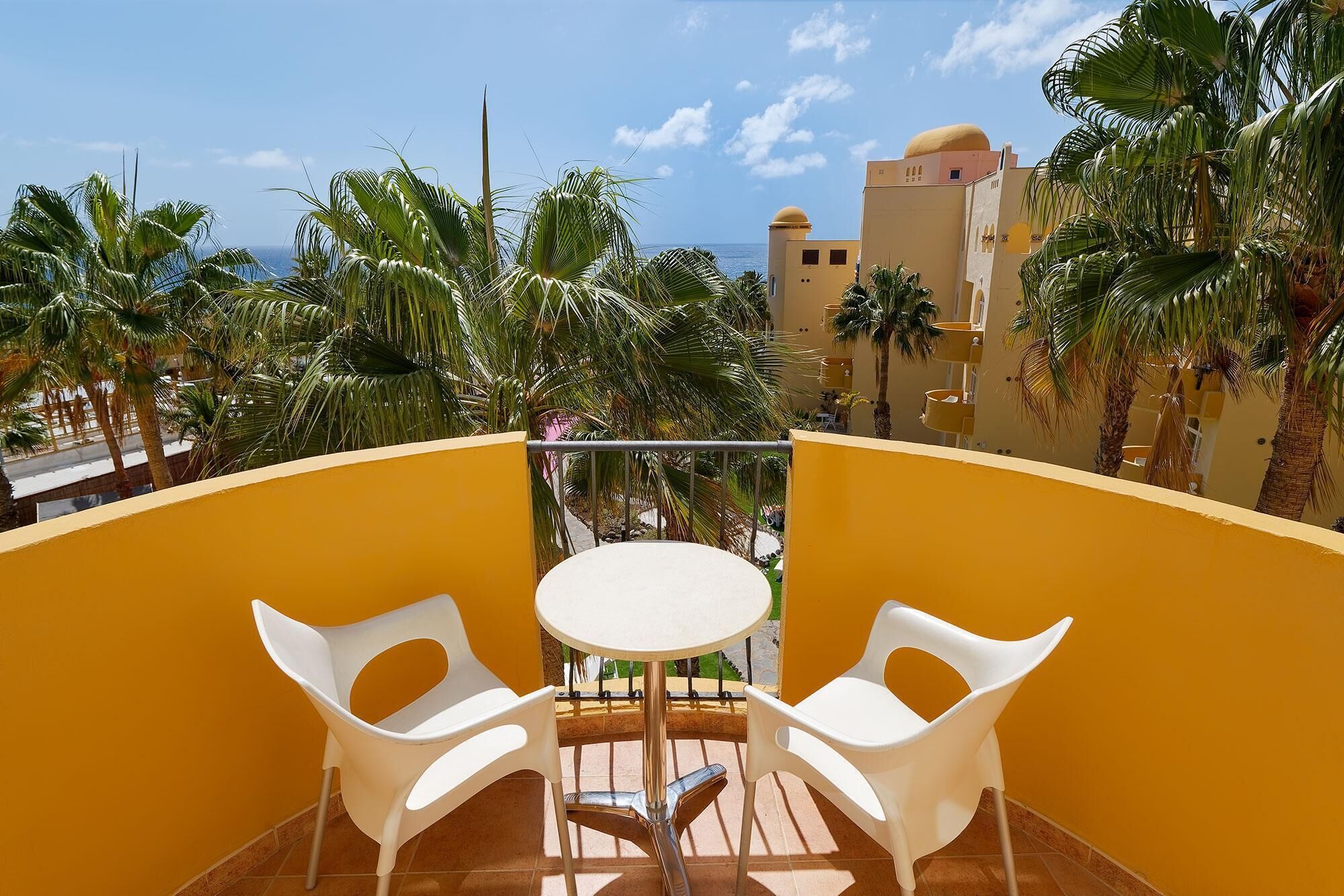 A balcony with two white plastic chairs and a small round table, overlooking palm trees and a building with a yellow dome.
