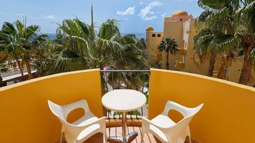 A balcony with two white plastic chairs and a small round table, overlooking palm trees and a building with a yellow dome.
