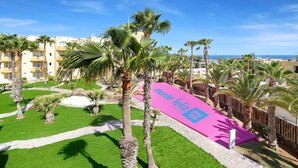 A resort courtyard with palm trees, a pink banner, and a view of the ocean. - Sport Hotel Monte Feliz - powered by Playitas (San Bartolomé de Tirajana)