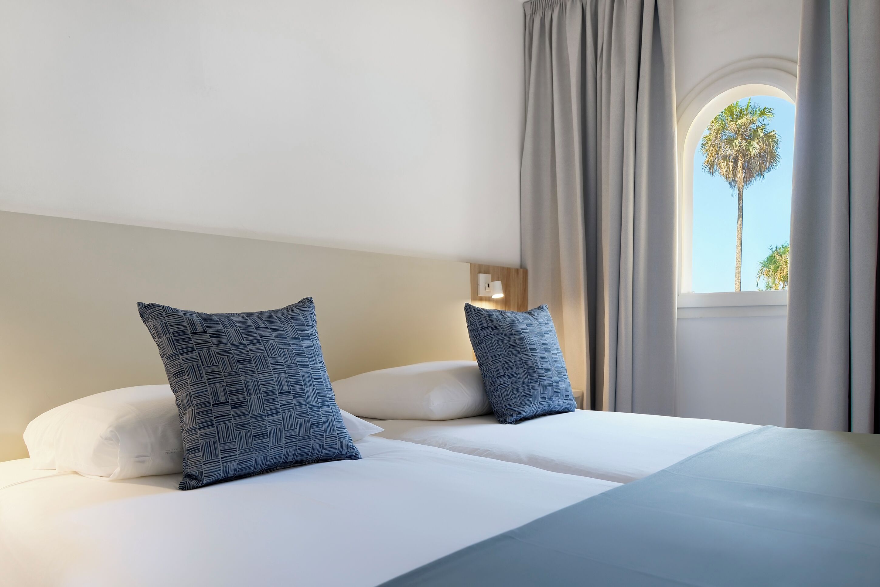A hotel room with two beds, a window with curtains, and a view of palm trees.
