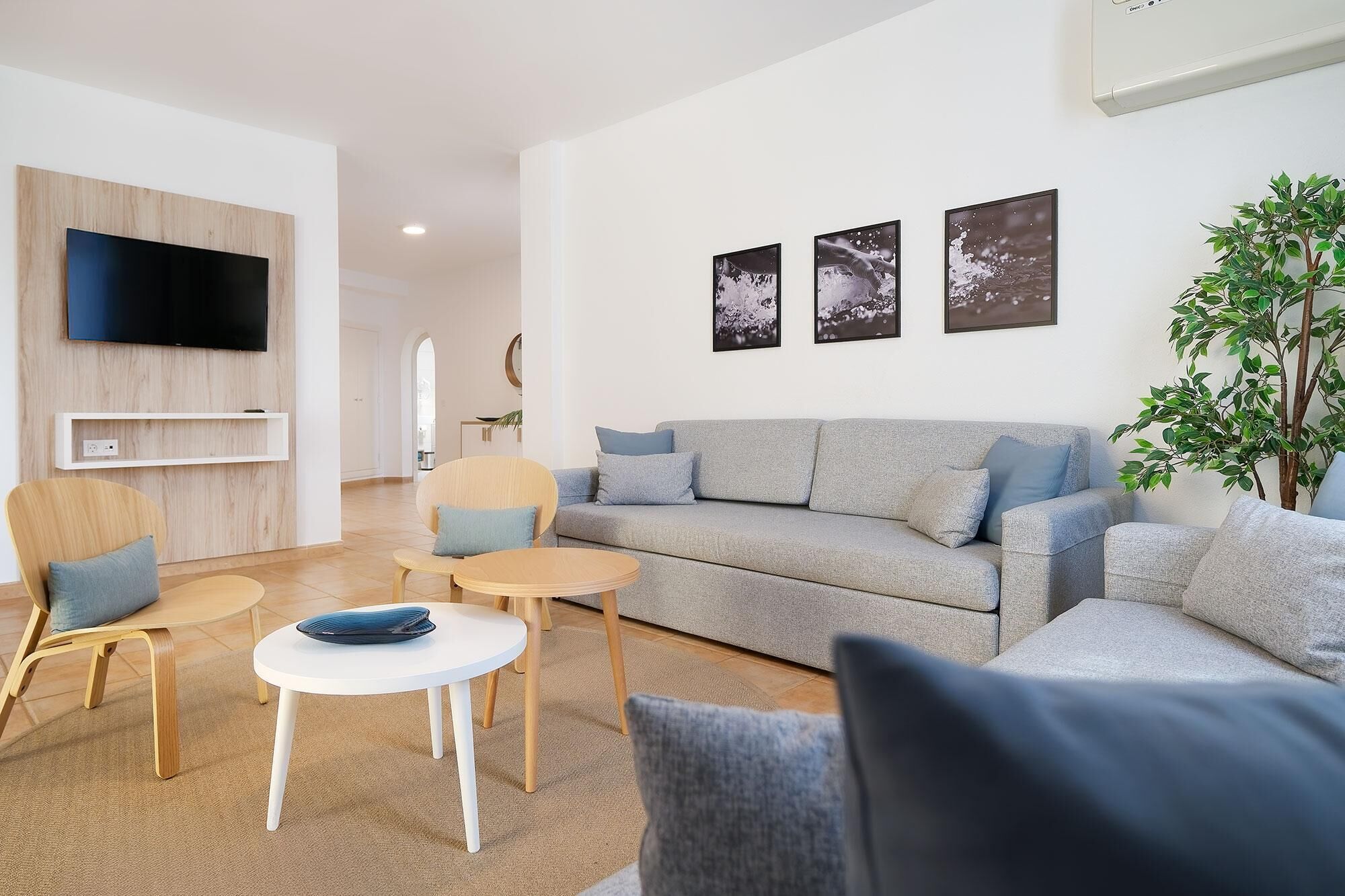 A modern living room with a grey sofa, two armchairs, a coffee table, and a flat-screen TV mounted on a wooden panel.