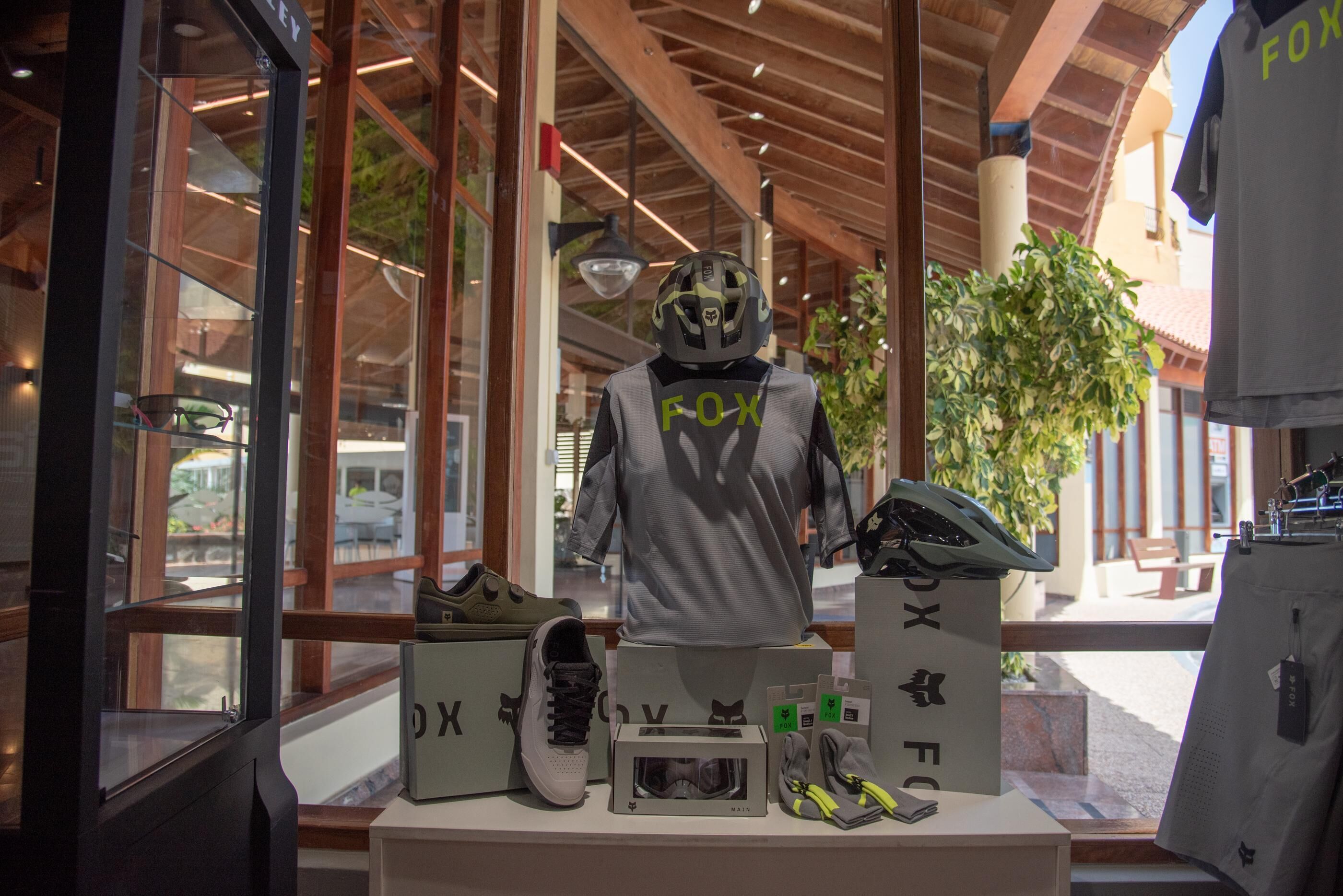 Un maniquí con casco y una camiseta con la palabra "FOX" en la espalda, frente a una exhibición de zapatos y accesorios.