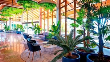 A spacious lobby with a high ceiling, large windows, and abundant indoor plants.