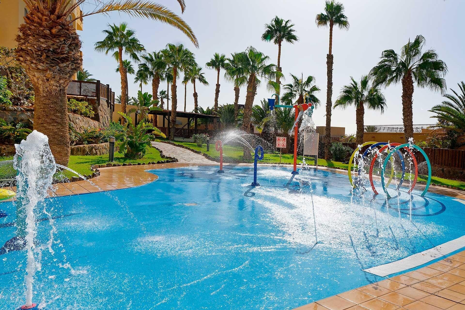 Piscina con chorros de agua, rodeada de palmeras y un sendero.