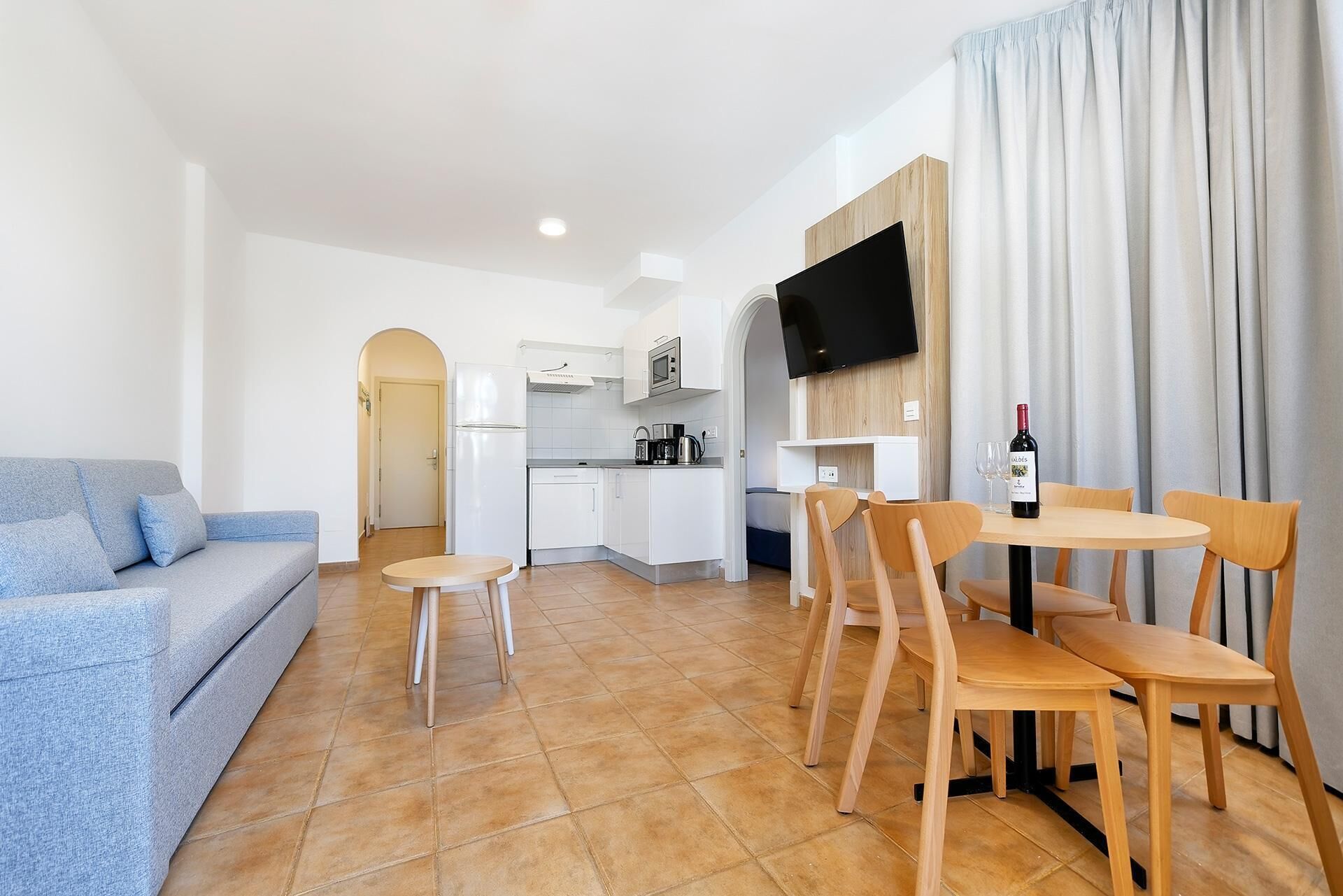 A compact living space with a kitchenette, a dining area with wooden chairs, a blue sofa, and a TV mounted on the wall.