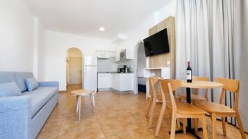 A compact living space with a kitchenette, a dining area with wooden chairs, a blue sofa, and a TV mounted on the wall.